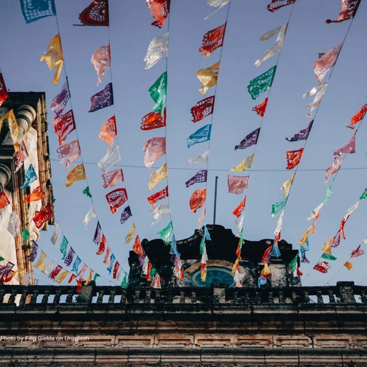 墨西哥旗幟橫幅五月五日慶祝背景 壁紙 — 色彩鮮豔的 papel picado 橫幅在晴朗的藍天下，在歷史悠久的石建築上方飄揚。