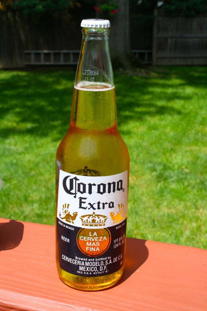 墨西哥科羅娜特級啤酒 壁紙 — 一瓶冰凍的Corona Extra啤酒瓶，帶有水珠，放在木桌上，背景是陽光明媚的綠色草坪。