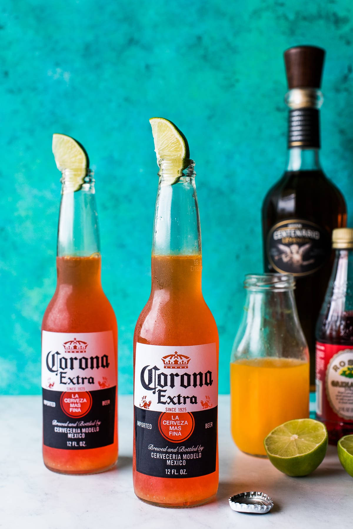 墨西哥紅色Corona Extra啤酒 壁紙 — 兩瓶冰凍的Corona Extra啤酒連同青檸角，旁邊放著一瓶龍舌蘭酒和新鮮的青檸半個，背景是充滿活力的藍綠色。