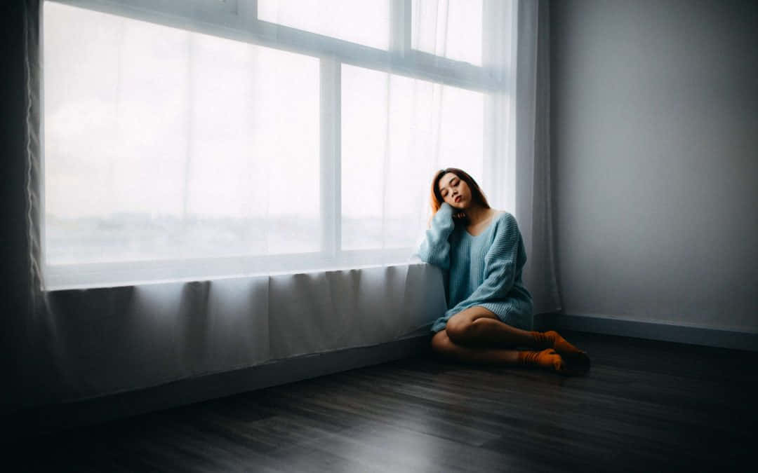 墮胎後掙扎的女性 壁紙 — 一位身穿藍色毛衣的女性靜靜地坐在木地板上，旁邊是一扇帶有薄紗窗簾的大窗戶，沐浴在柔和自然光中。