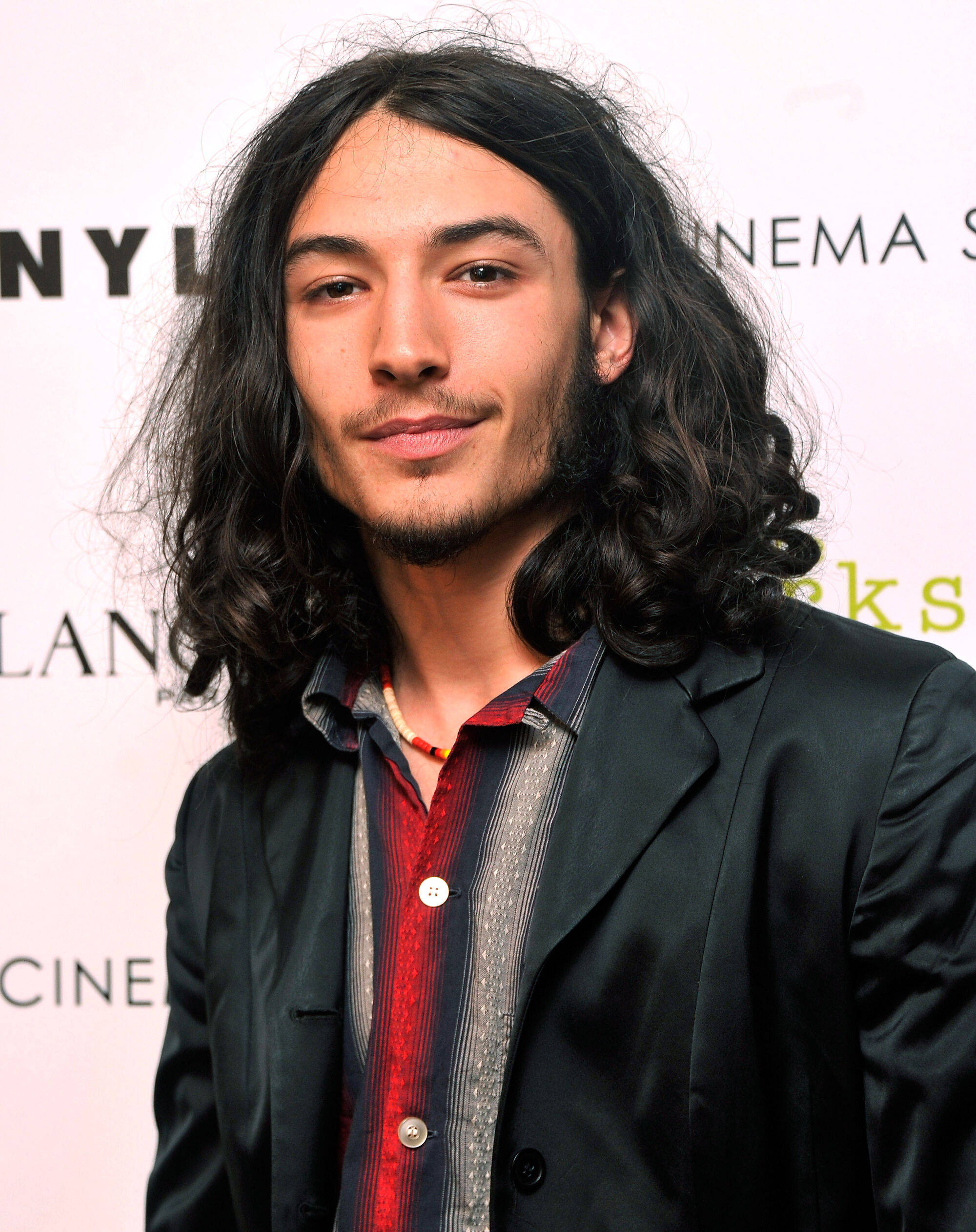 《壁花少年》電影海報：Ezra Miller 壁紙 — 一位留著深色長捲髮和鬍鬚的男子在電影活動中穿著黑色西裝外套內搭條紋襯衫，對著鏡頭微笑。