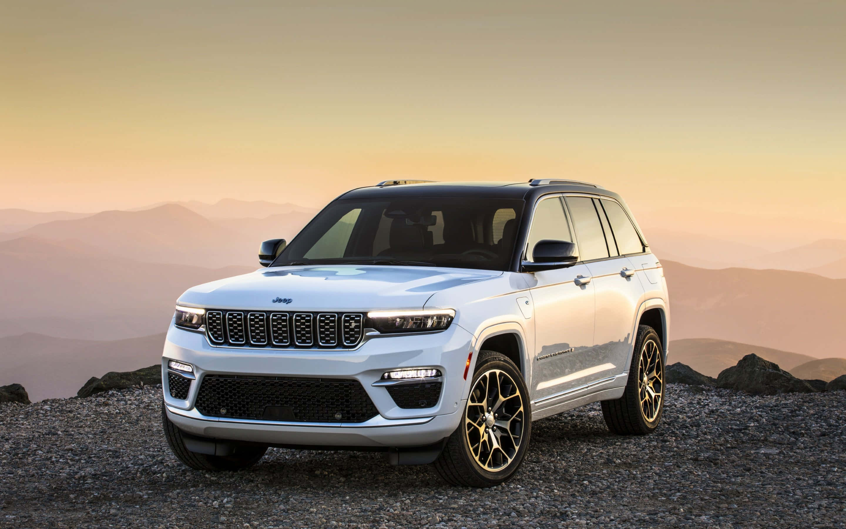 壯觀吉普瓦格尼爾駛過風景公路 壁紙 — 一架時尚的白色Jeep Grand Cherokee停泊在日落時分的岩石山脊上，背景是層層疊疊的山脈。