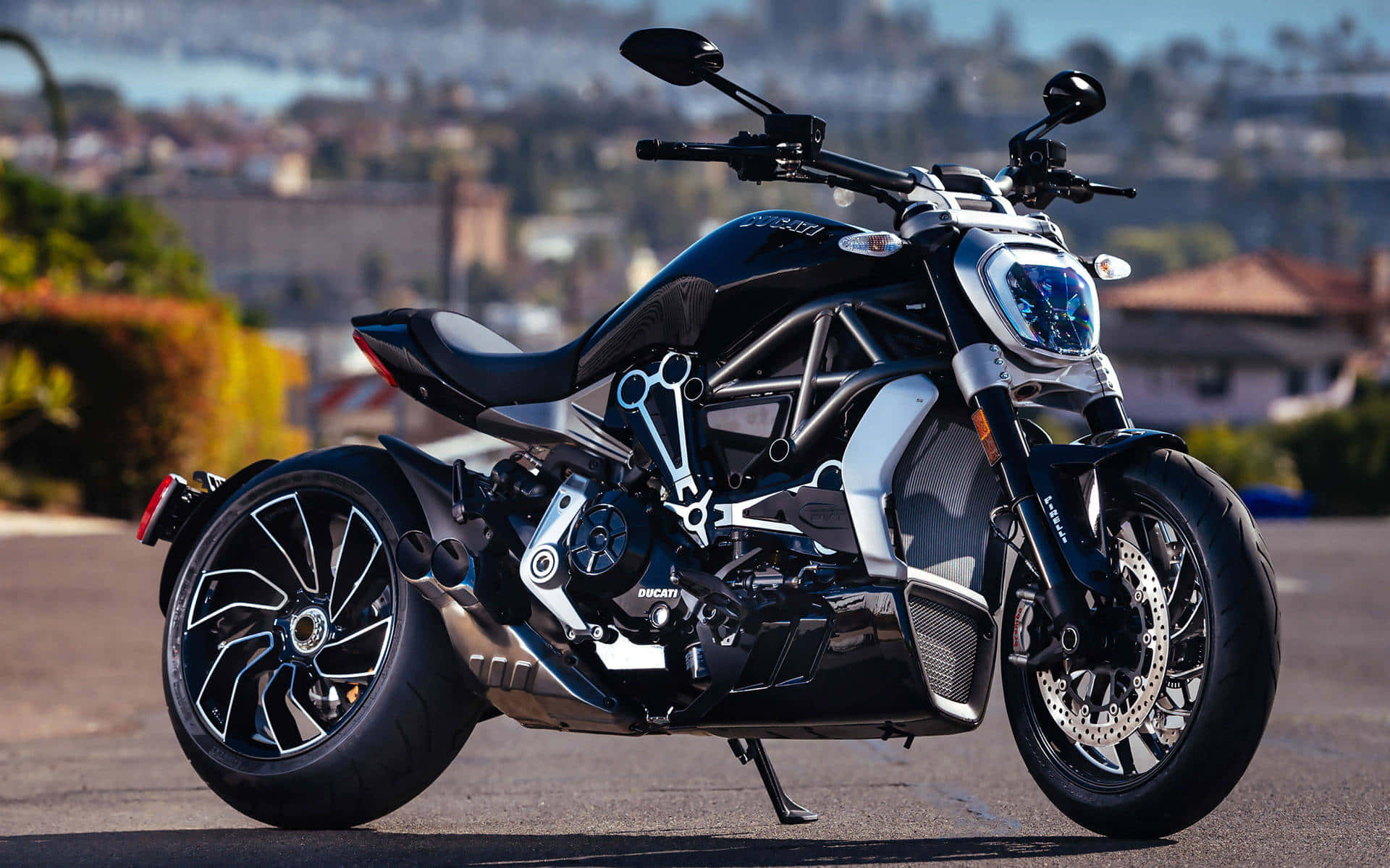 壯觀的杜卡迪XDiavel機車背景 壁紙 — 一輛時尚的黑色Ducati電單車，帶有銀色點綴，停泊在陽光明媚的道路上，俯瞰著朦朧的海岸小鎮。