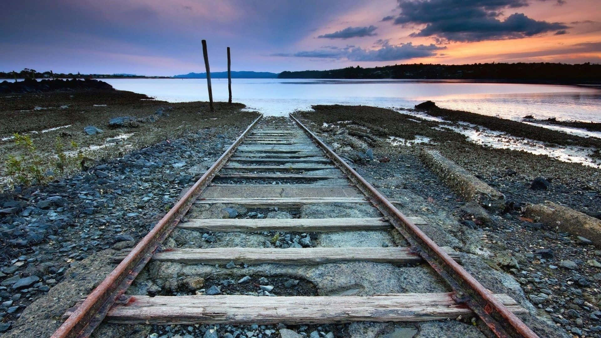 壯麗山脈日出Full HD 壁紙 — 廢棄的鐵路軌道在戲劇性的暮色天空下延伸向平靜的水面地平線。