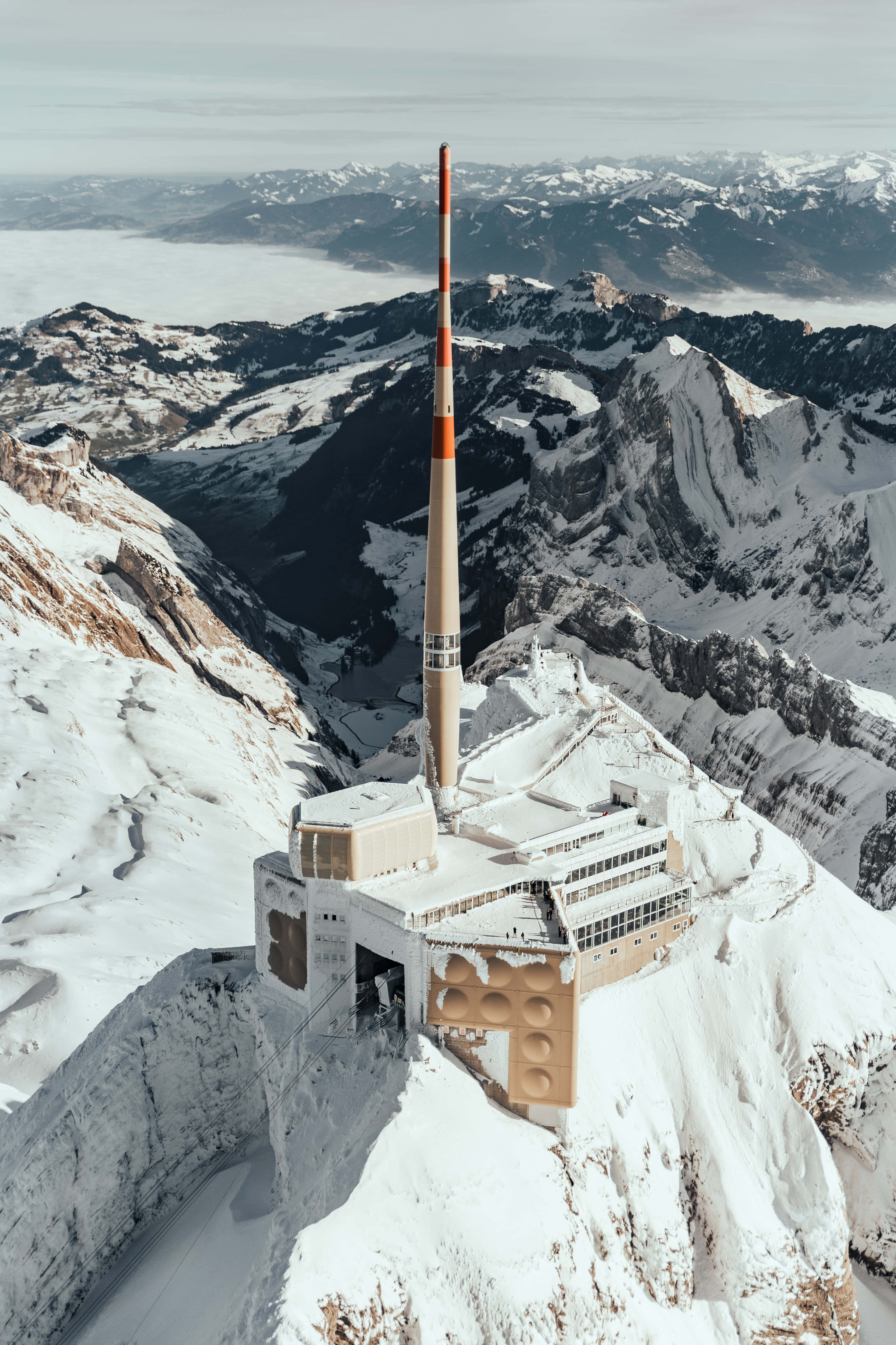 壯麗山脈日落風景 壁紙 — 一個積雪的山頂上有一座現代建築，帶有高大的紅白相間塔樓，俯瞰廣闊的阿爾卑斯山谷。