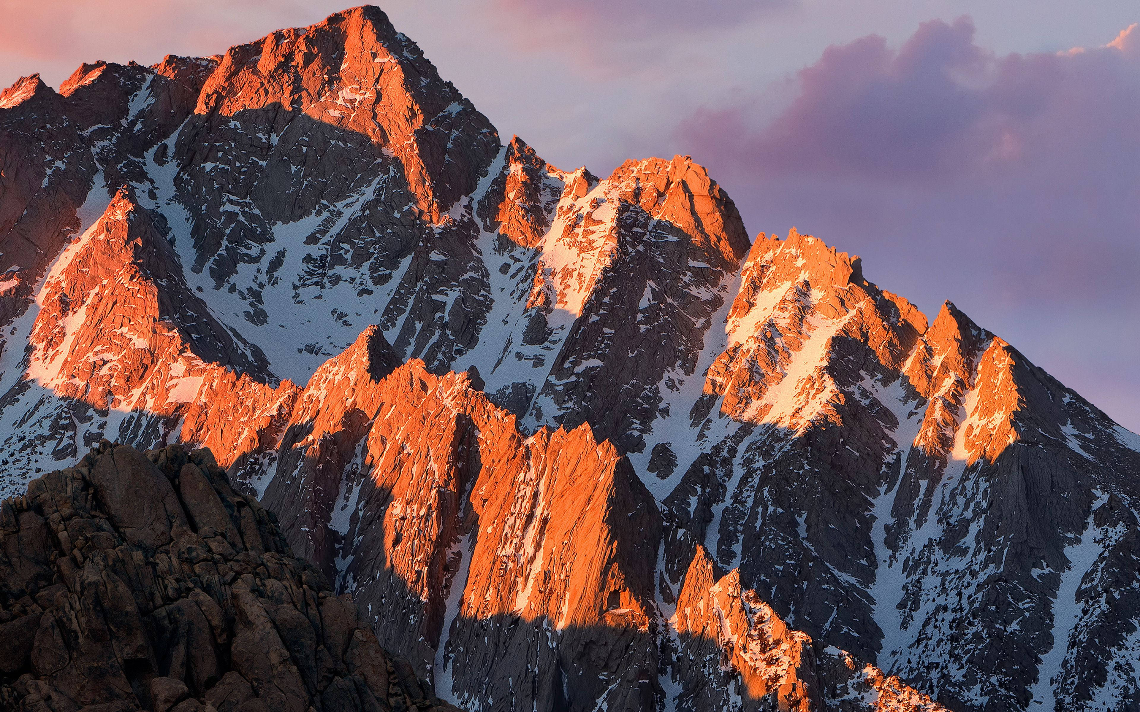 壯麗山脊 macOS 壁紙 — 夕陽將鋸齒狀的雪山山峰沐浴在溫暖的橙色光芒中，映襯著柔和的紫色天空。