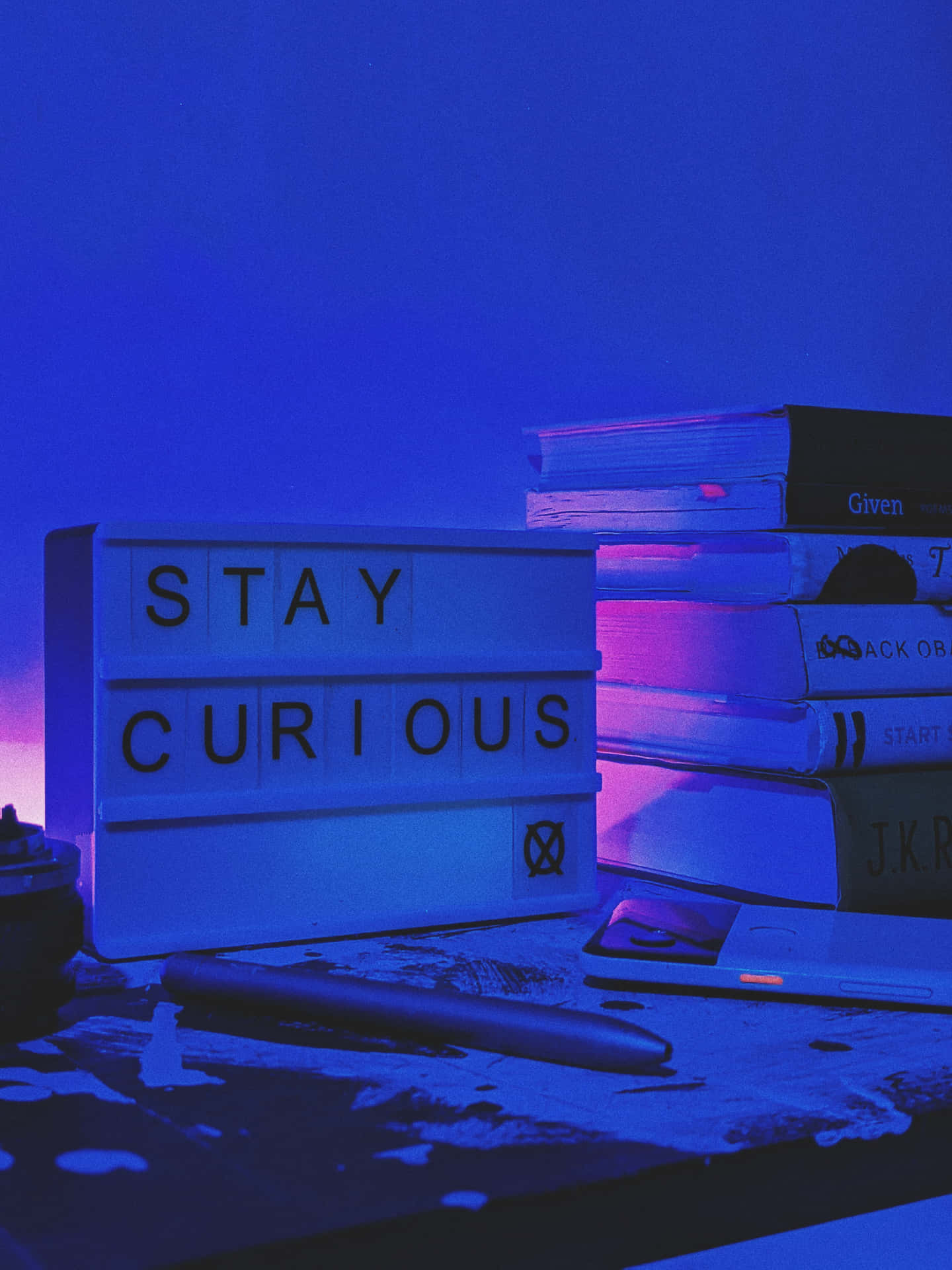 一個寫著「STAY CURIOUS」的燈箱，旁邊有書籍和一支筆，桌上濺滿油漆，在霓虹光下。