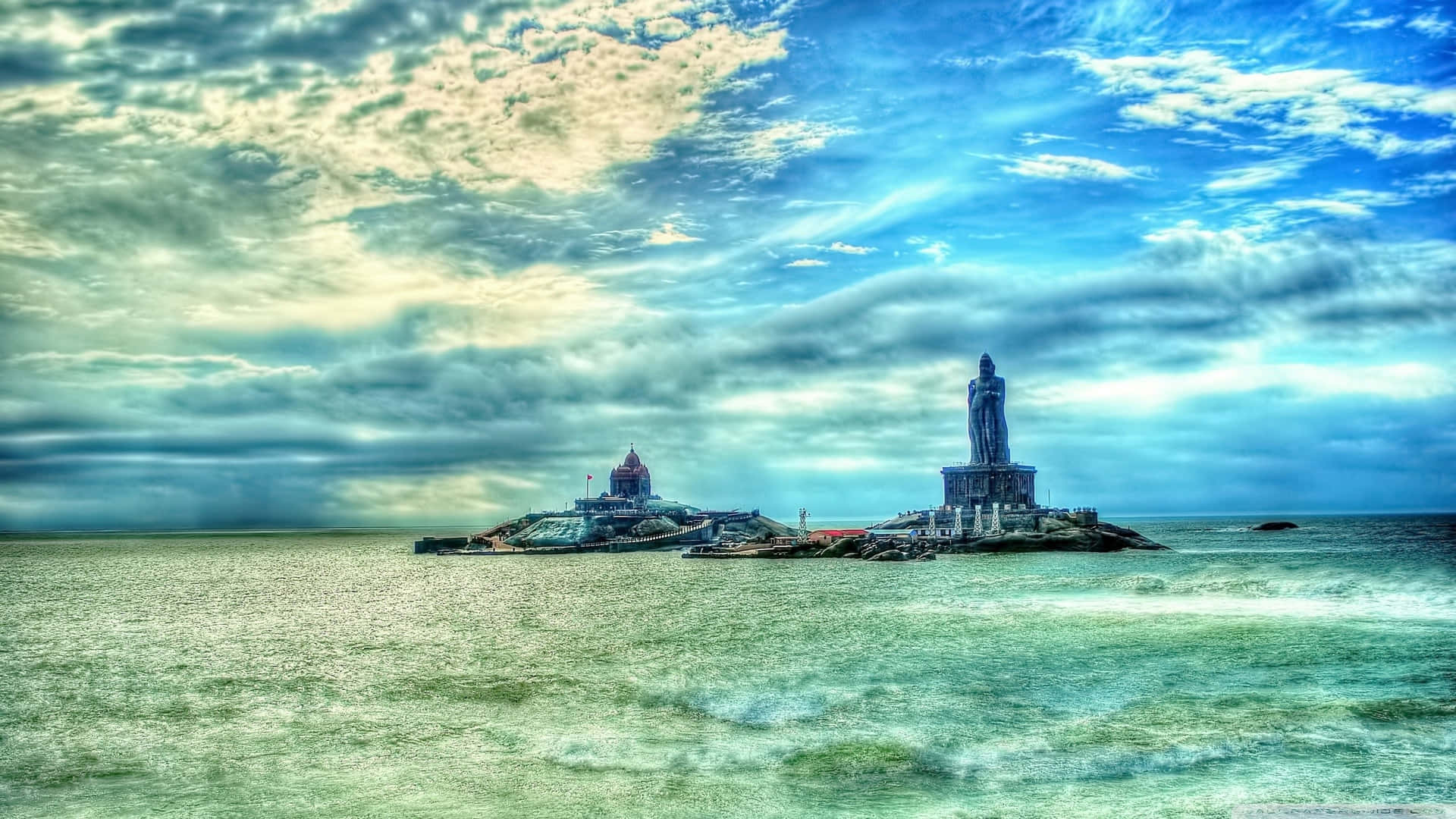 在多雲的天空下，海中的岩石島上矗立著一座高聳的雕像和寺廟。