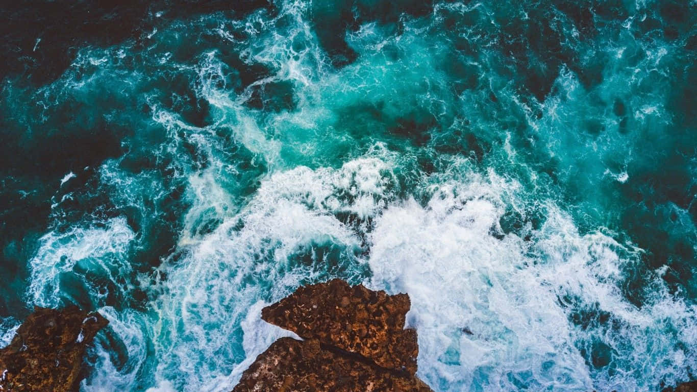 壯麗海洋日落 壁紙 — 從高空俯瞰，碧綠色的海浪拍打著崎嶇的棕色岩石，激起白色泡沫。