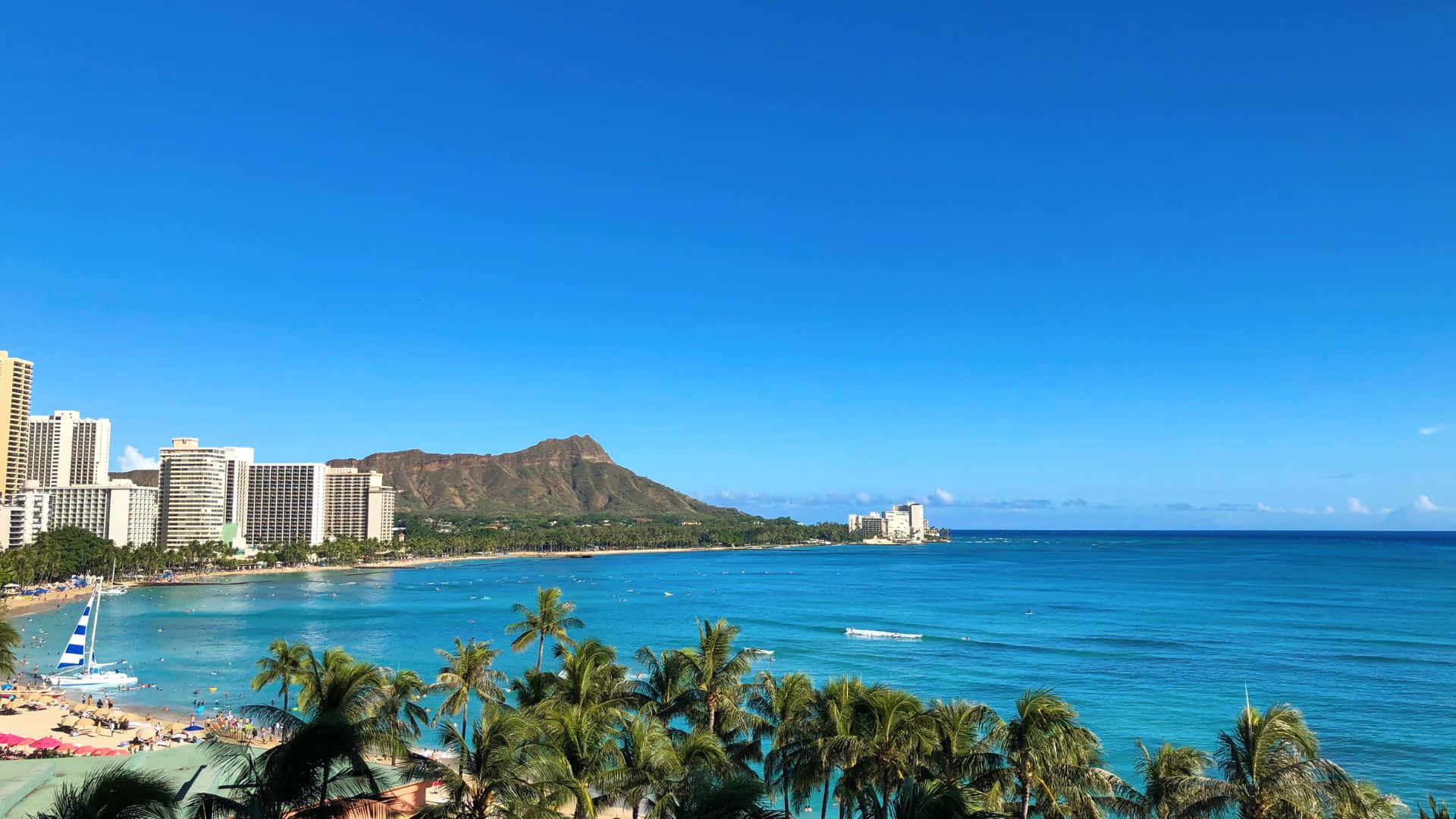 壯麗的夏威夷日落 壁紙 — 充滿活力的熱帶海灘場景，有碧綠的海水、棕櫚樹和一座標誌性的火山，在晴朗的藍天下聳立在現代酒店後面。