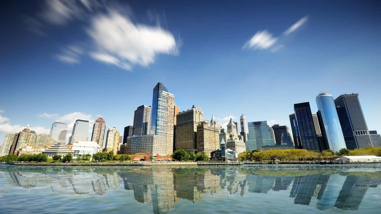 壯麗的紐約市 壁紙 — 充滿活力的城市天際線，高聳的摩天大樓倒映在平靜的水面上，天空湛藍，雲彩斑駁。