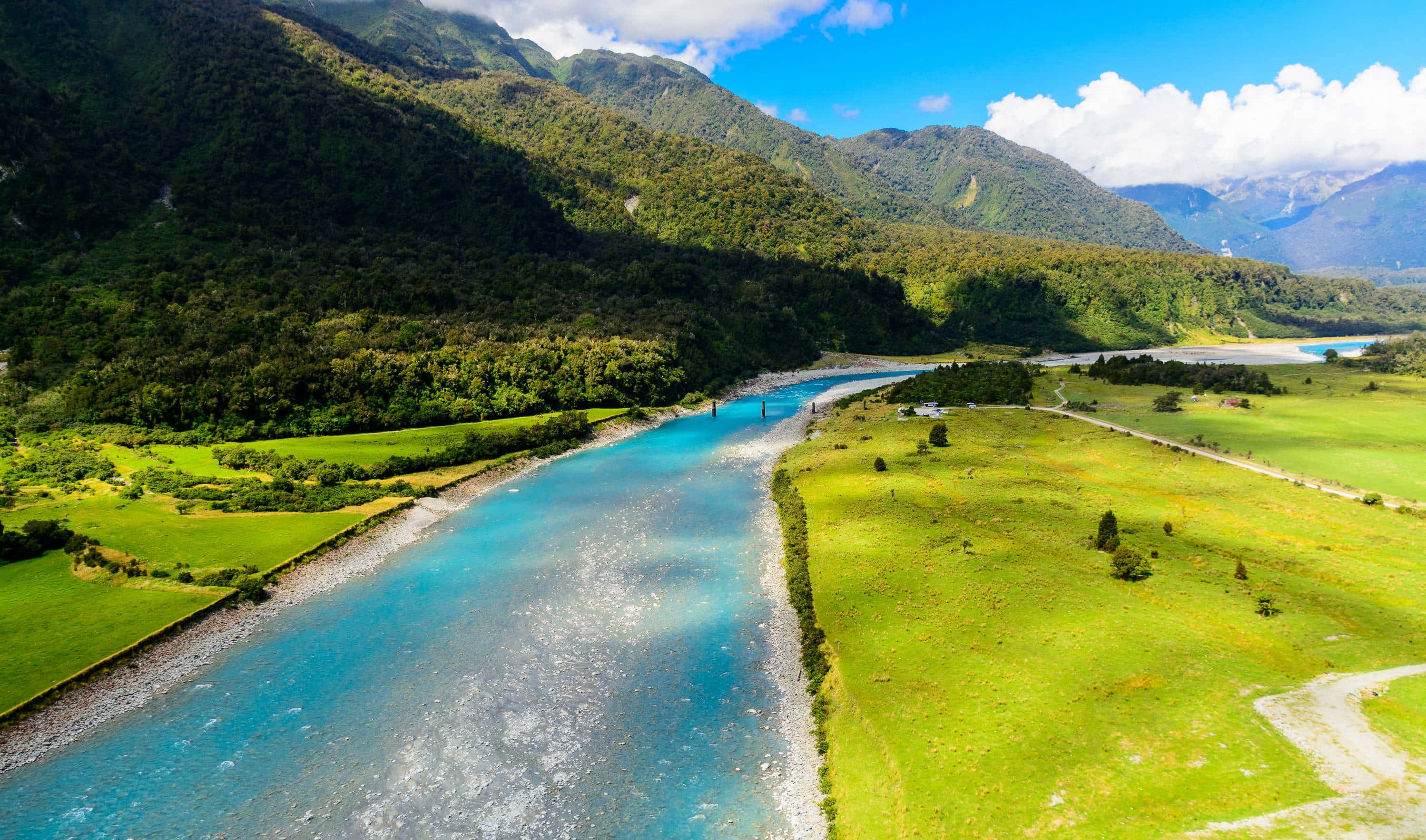壯麗的自然風景 壁紙 — 從高空俯瞰，一條充滿活力的藍綠色河流蜿蜒穿過茂密的綠色山谷，兩旁是森林覆蓋的山脈，上方是明亮的藍天。
