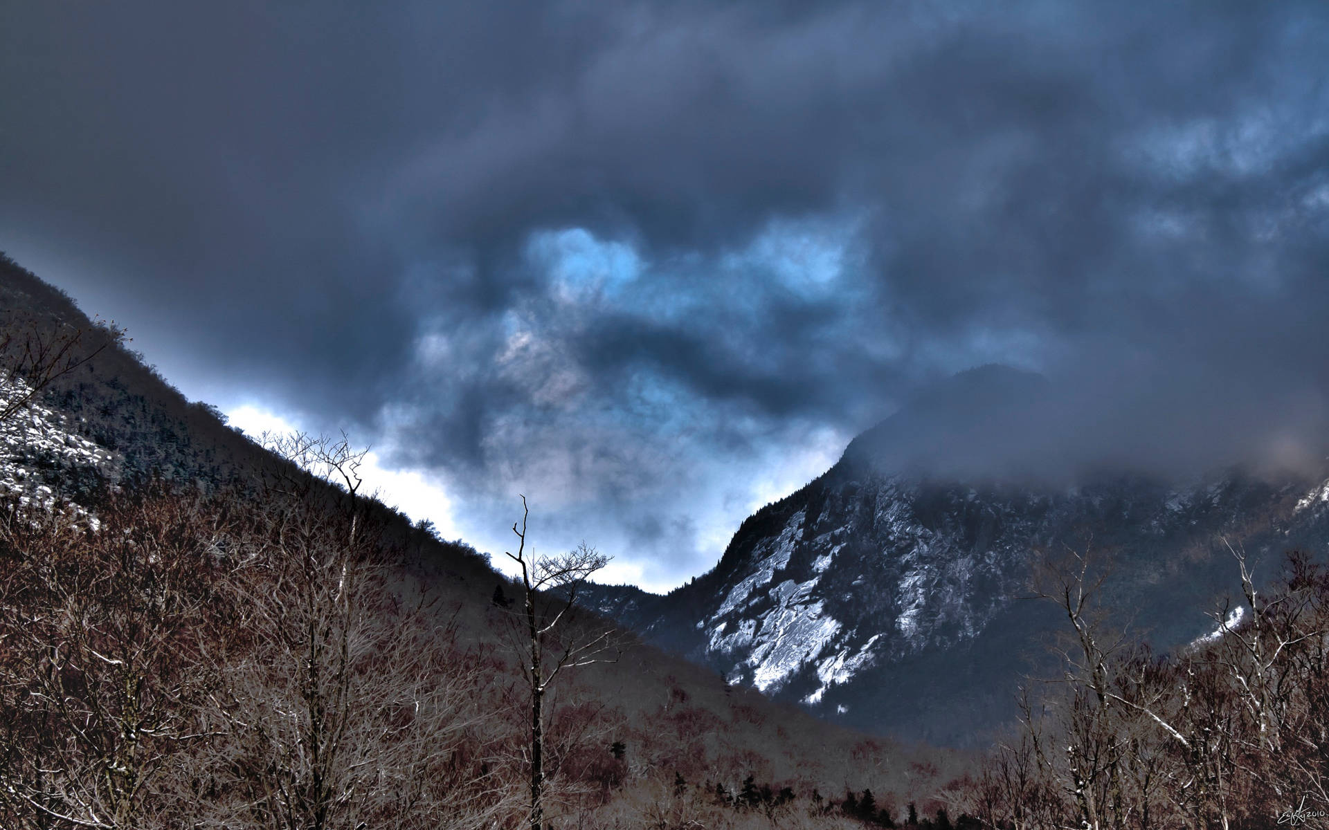 冬季山脈，有雪和光禿禿的樹木，在暴風雨的天空下，帶有藍色斑塊。