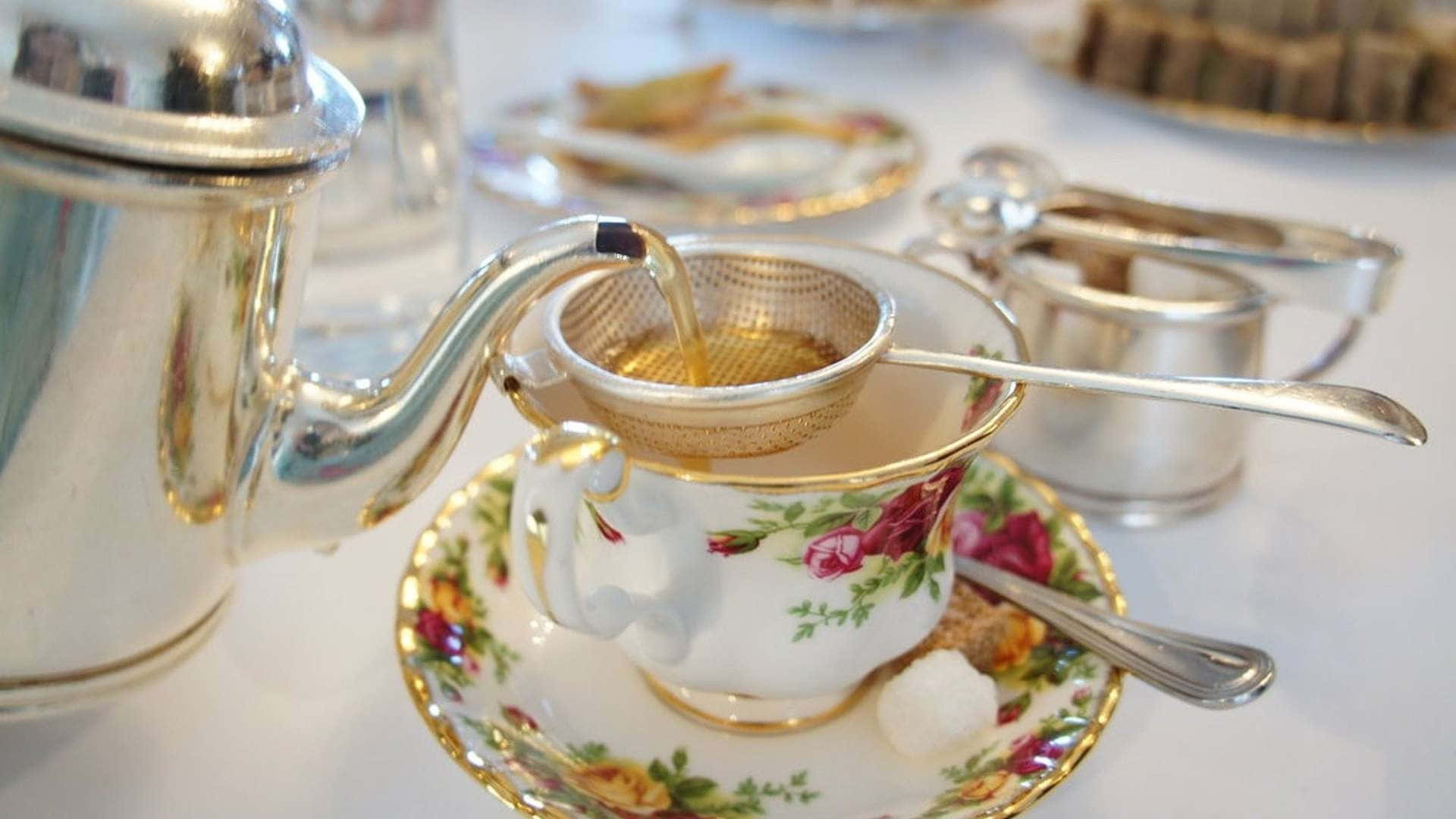 壺倒茶入杯圖 壁紙 — 金色茶湯透過濾網倒入花卉茶杯，旁邊有銀器和方糖。