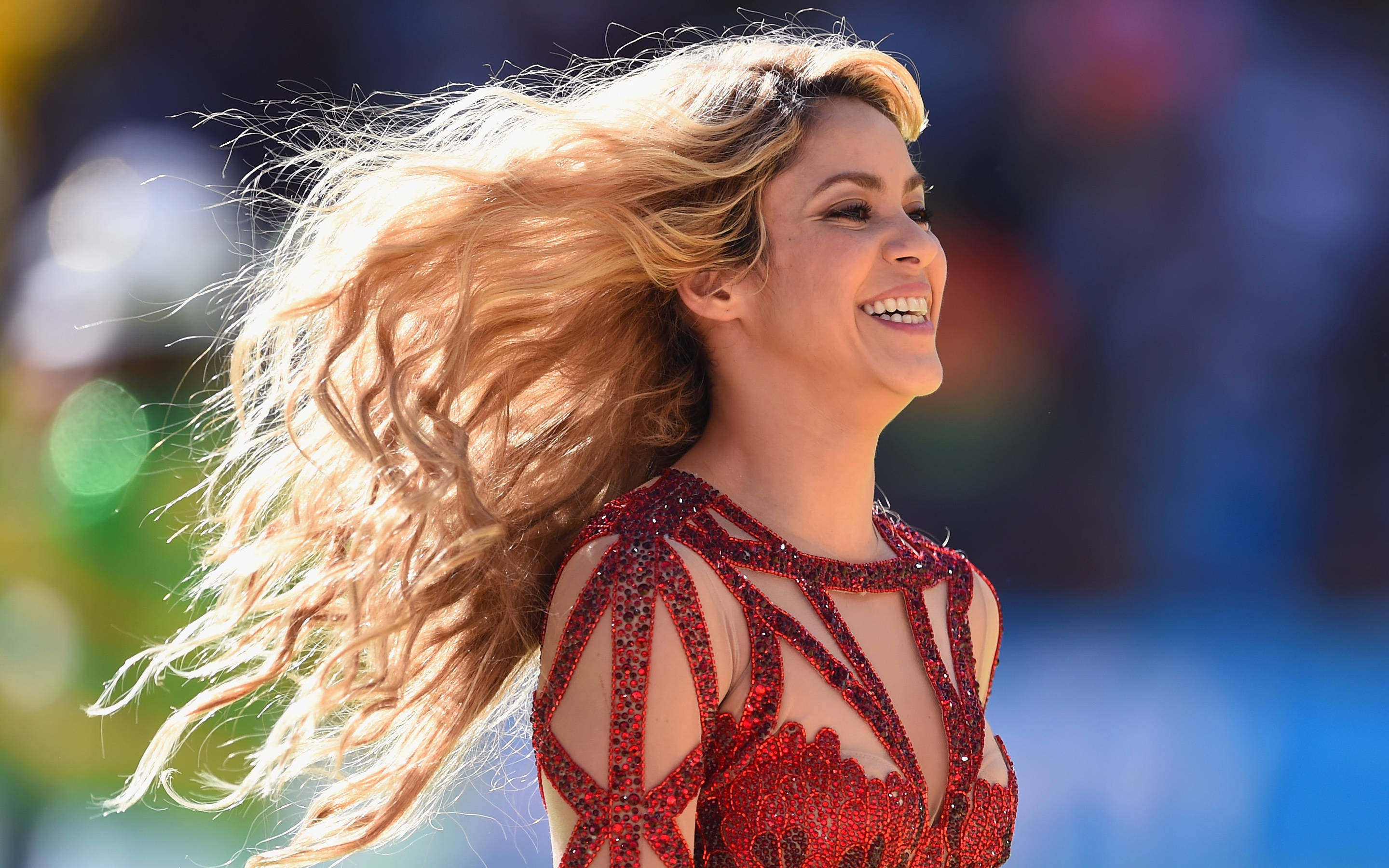 夏奇拉2020超級盃演出 壁紙 — Shakira 在模糊的體育場背景下，飄逸的金色頭髮和閃閃發光的紅色服裝，露出燦爛的笑容。