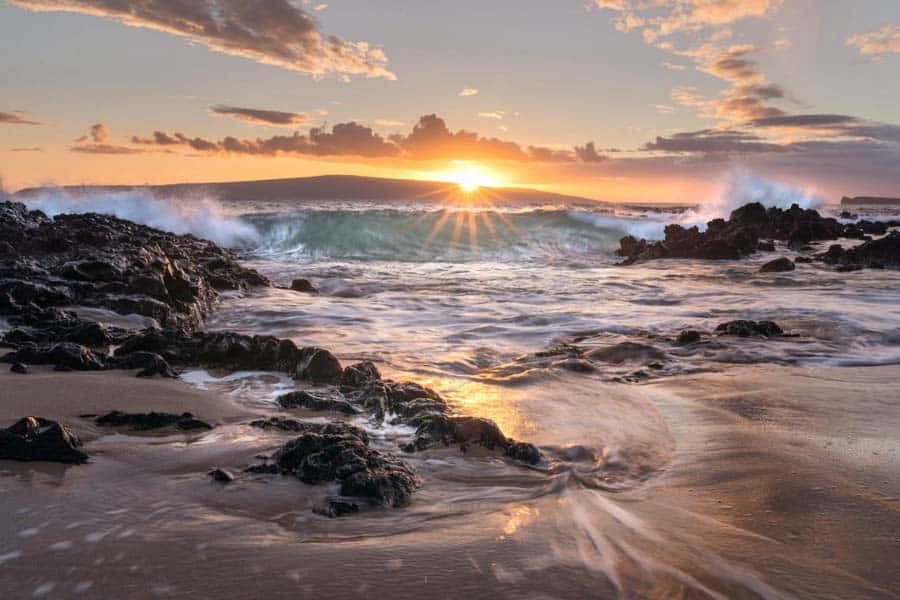 夏威夷日落岩石海岸圖 壁紙 — 夕陽穿透雲層，海浪拍打沙灘上的深色火山岩，水花四濺。