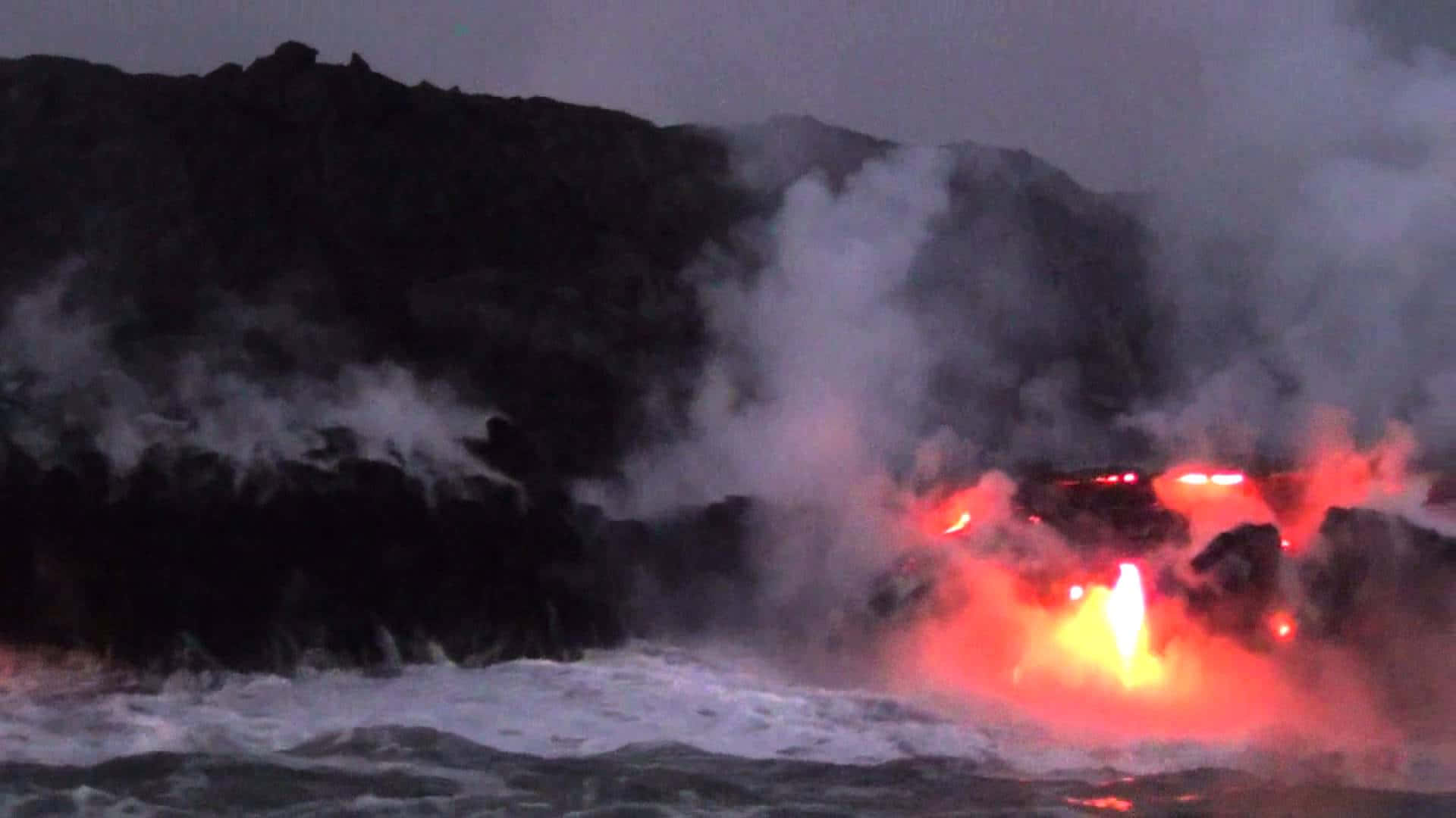 夏威夷火山國家公園基拉韋厄火山 壁紙 — 熔岩猛烈地噴發入海，在黑暗的火山岩和翻騰的蒸汽上投射出熾熱的紅光。