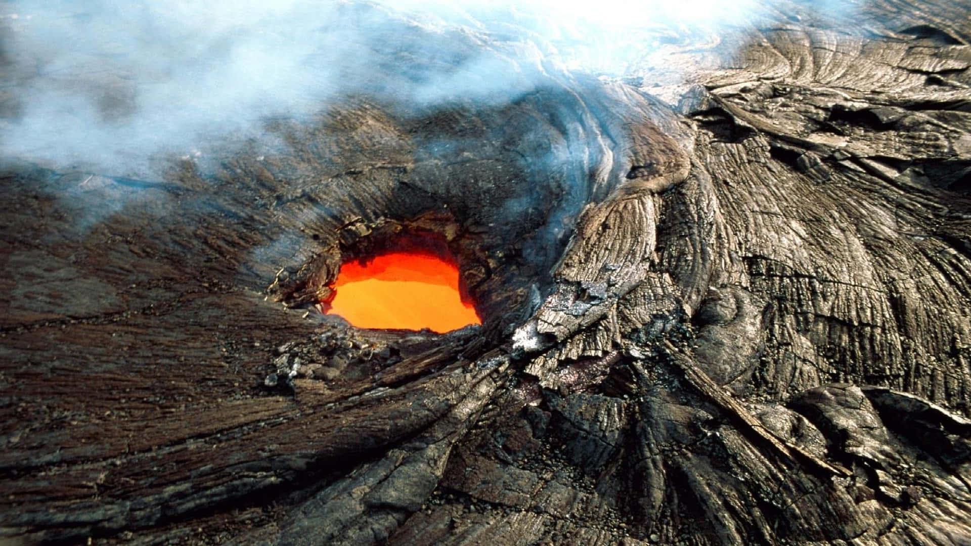 夏威夷火山國家公園洛伊希海山 壁紙 — 一個火山口噴發出熾熱的橙色熔岩，周圍環繞著崎嶇的黑色岩石和滾滾白煙。