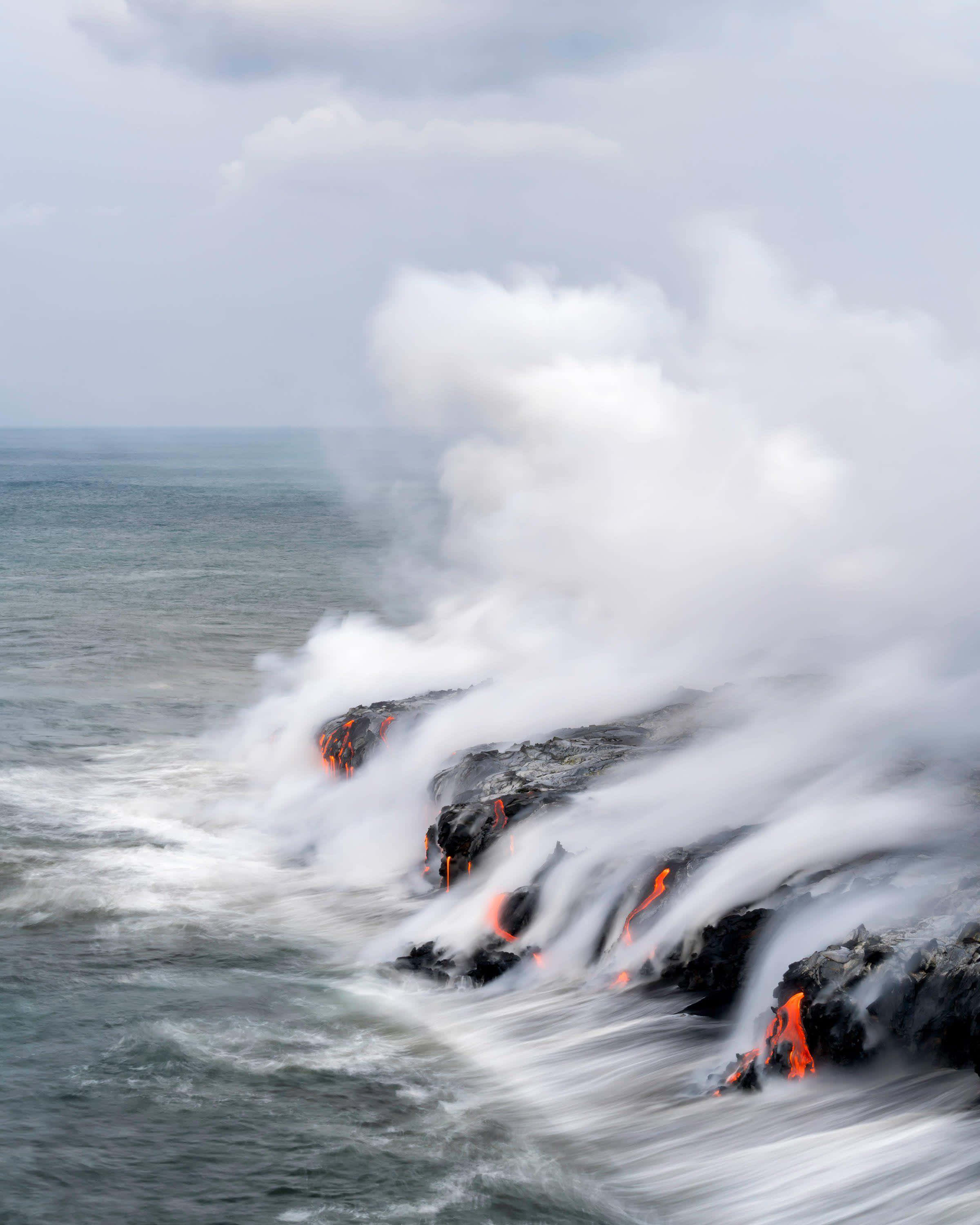 夏威夷火山國家公園熔岩湧入海洋 壁紙 — 熔岩流與海浪相遇，在黑色岩石上形成翻滾的蒸氣和發光的橙色裂縫。