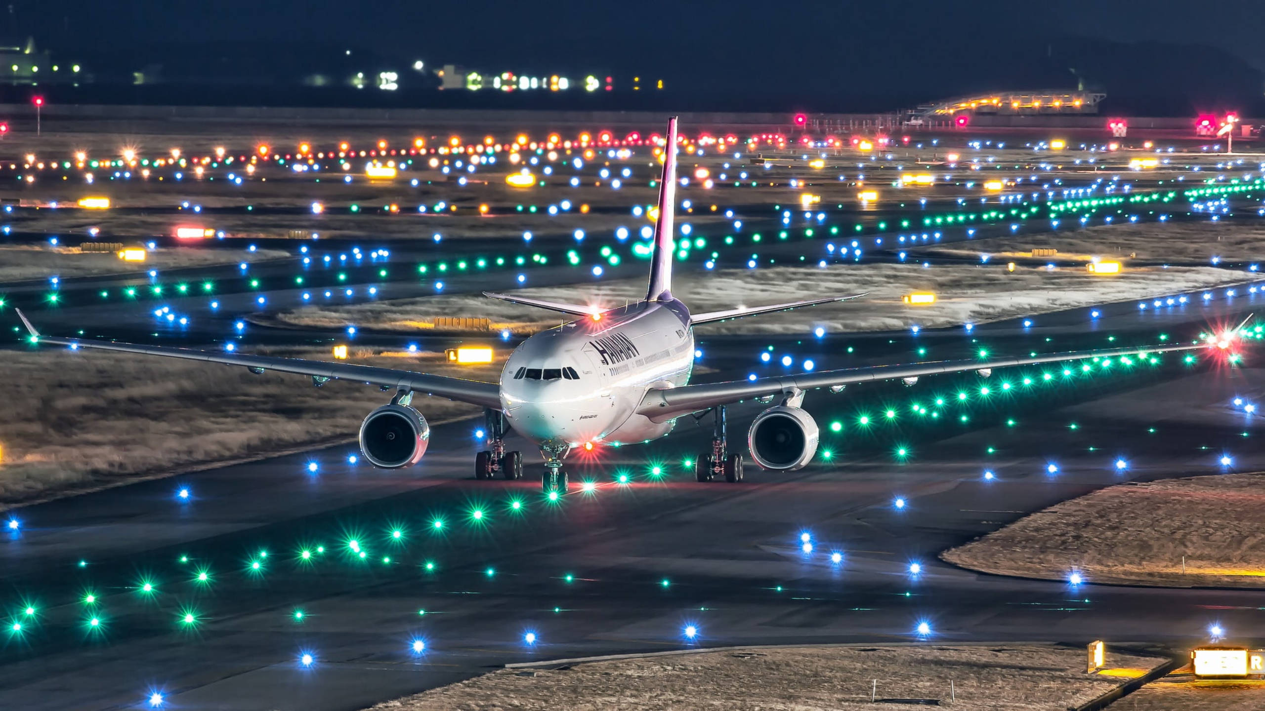 夏威夷航空飛機在跑道上 壁紙 — 一架大型飛機在夜間於一條被鮮豔綠色和藍色燈光照亮的跑道上滑行，遠處的機場建築物在背景中發光。