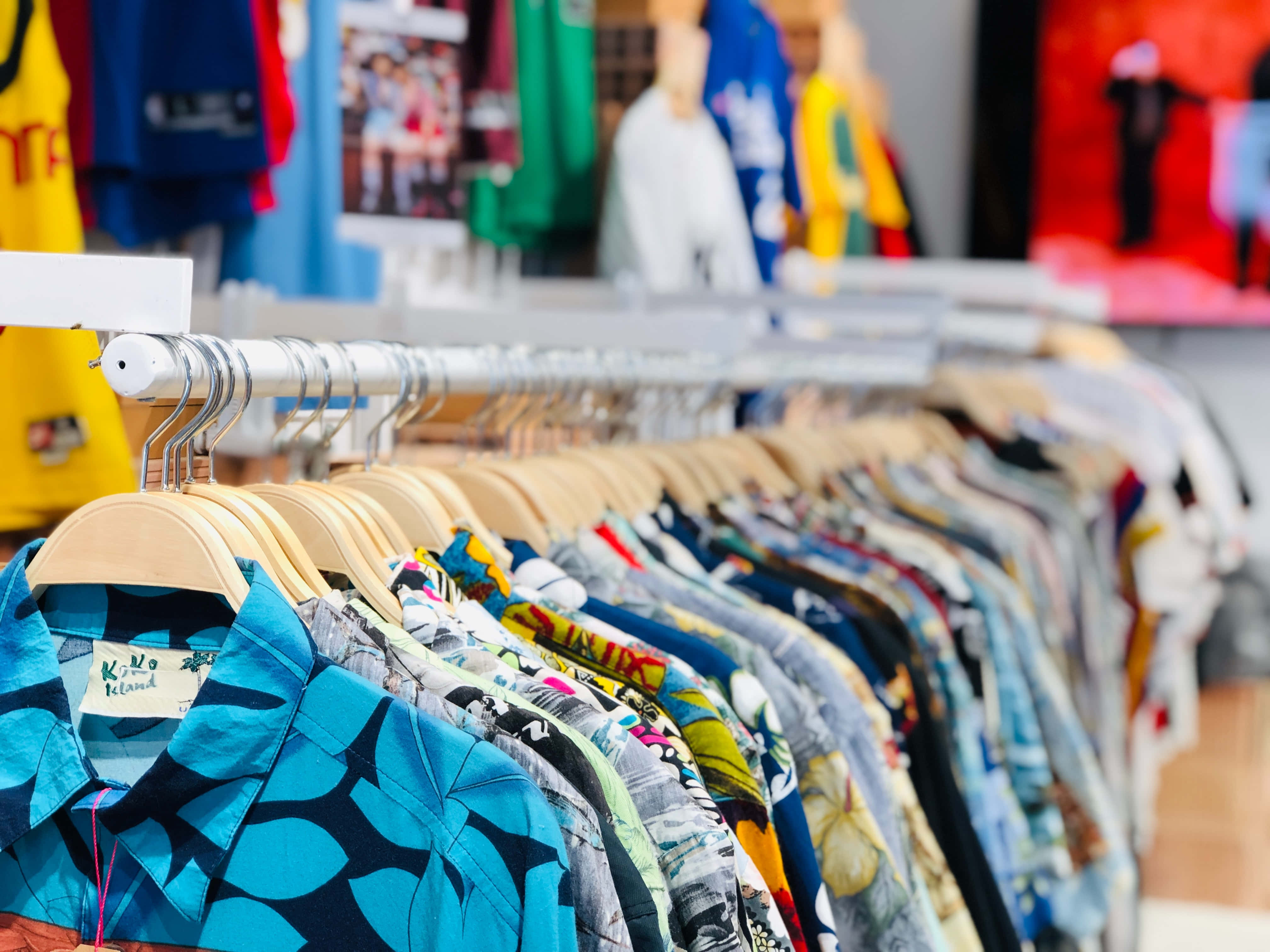 夏季POLO衫服裝店畫面 壁紙 — 一排色彩繽紛的夏威夷恤衫整齊地掛在明亮零售店的木衣架上。