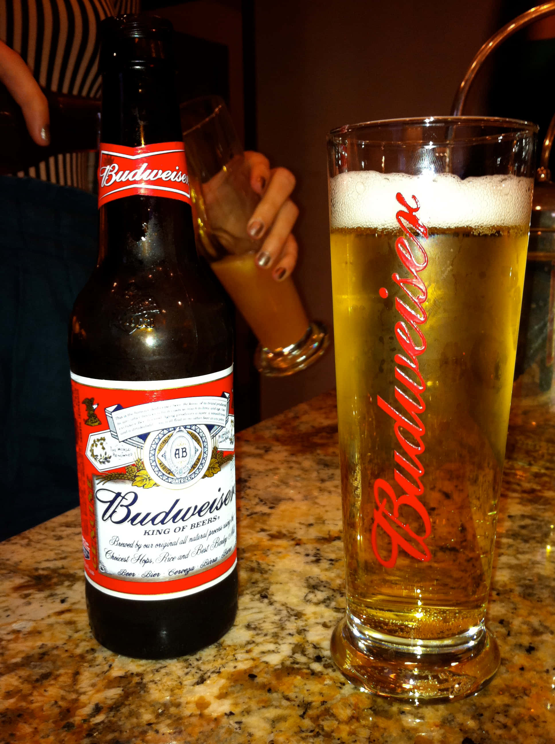 夏日享受冰涼啤酒 壁紙 — 一瓶冰凍的Budweiser啤酒和一個玻璃杯放在有斑點的酒吧櫃檯上，背景中有一隻手拿著另一個玻璃杯。
