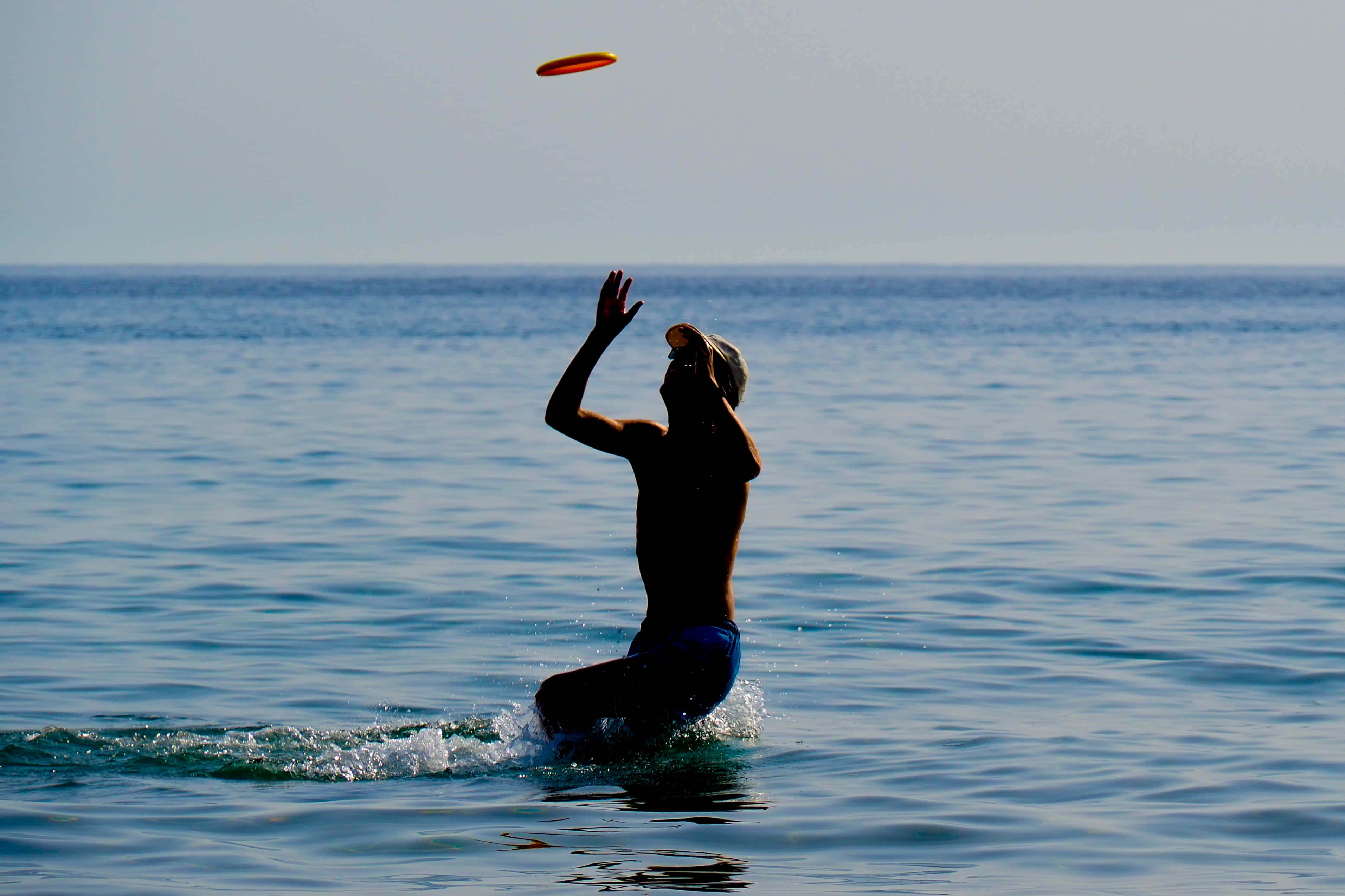 夏日季節 1080p 終極飛盤背景 壁紙 — 一個剪影人物從海浪中躍起，伸手去接在明亮日光下飛行的橙色飛盤。