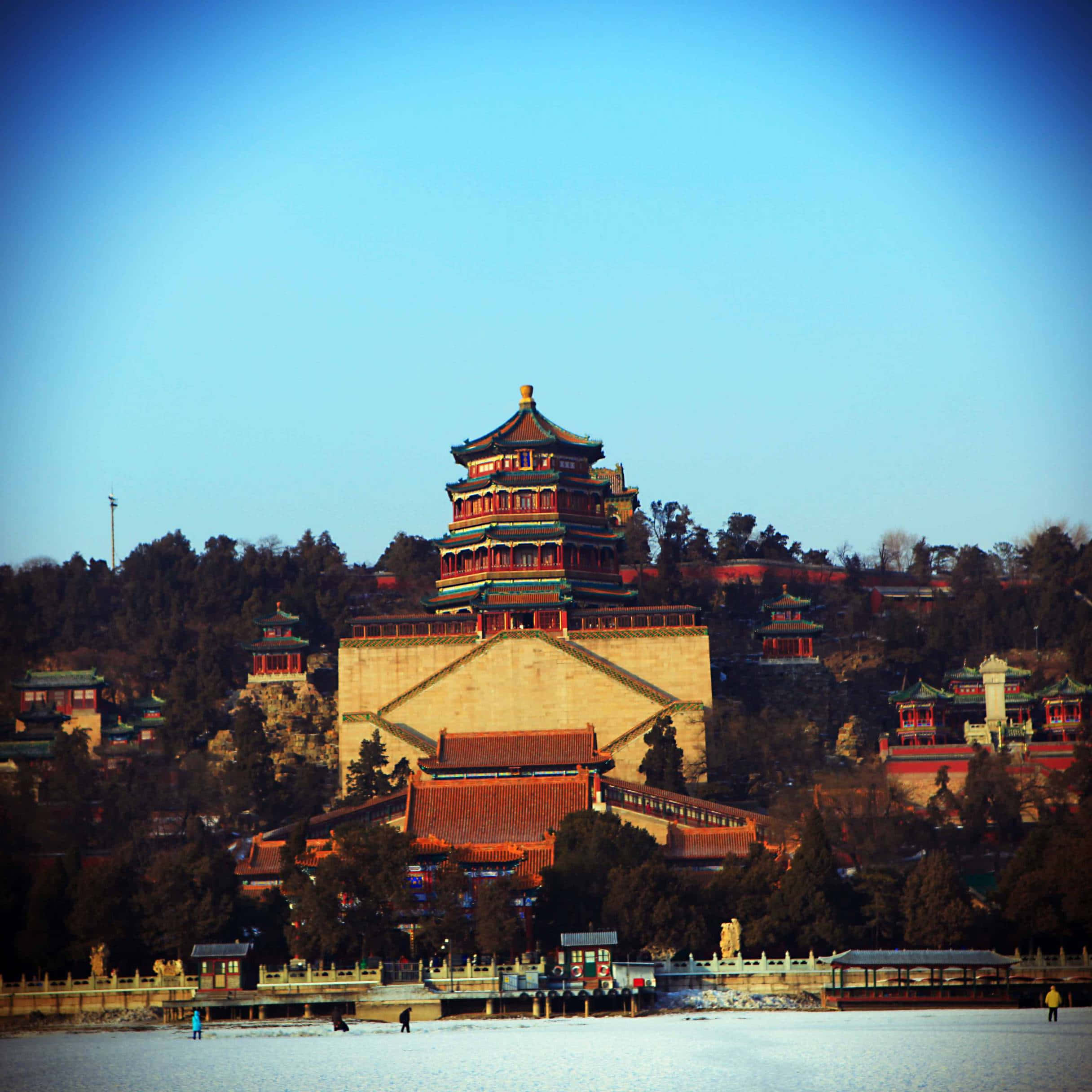 夏日宮殿全景 壁紙 — 一座雄偉的傳統中式寶塔從結冰的湖面升起，在清澈的藍天下，樹木稀疏。
