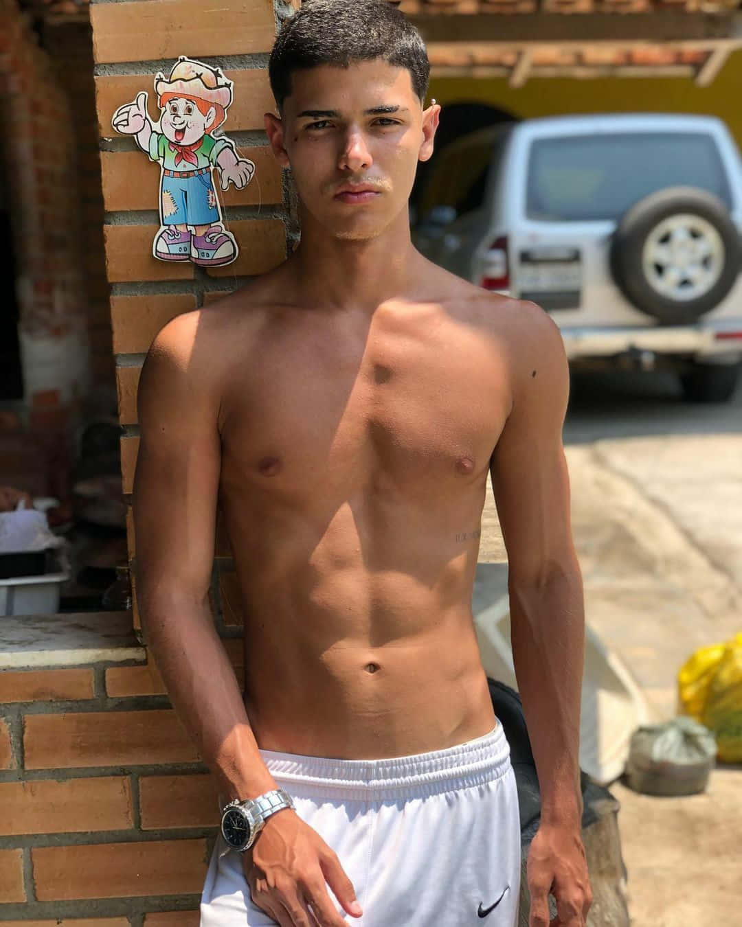 夏日帥氣男孩照片 壁紙 — 一個赤膊上身、腹肌線條分明的年輕男子，在明亮陽光下自信地靠著磚牆站立。