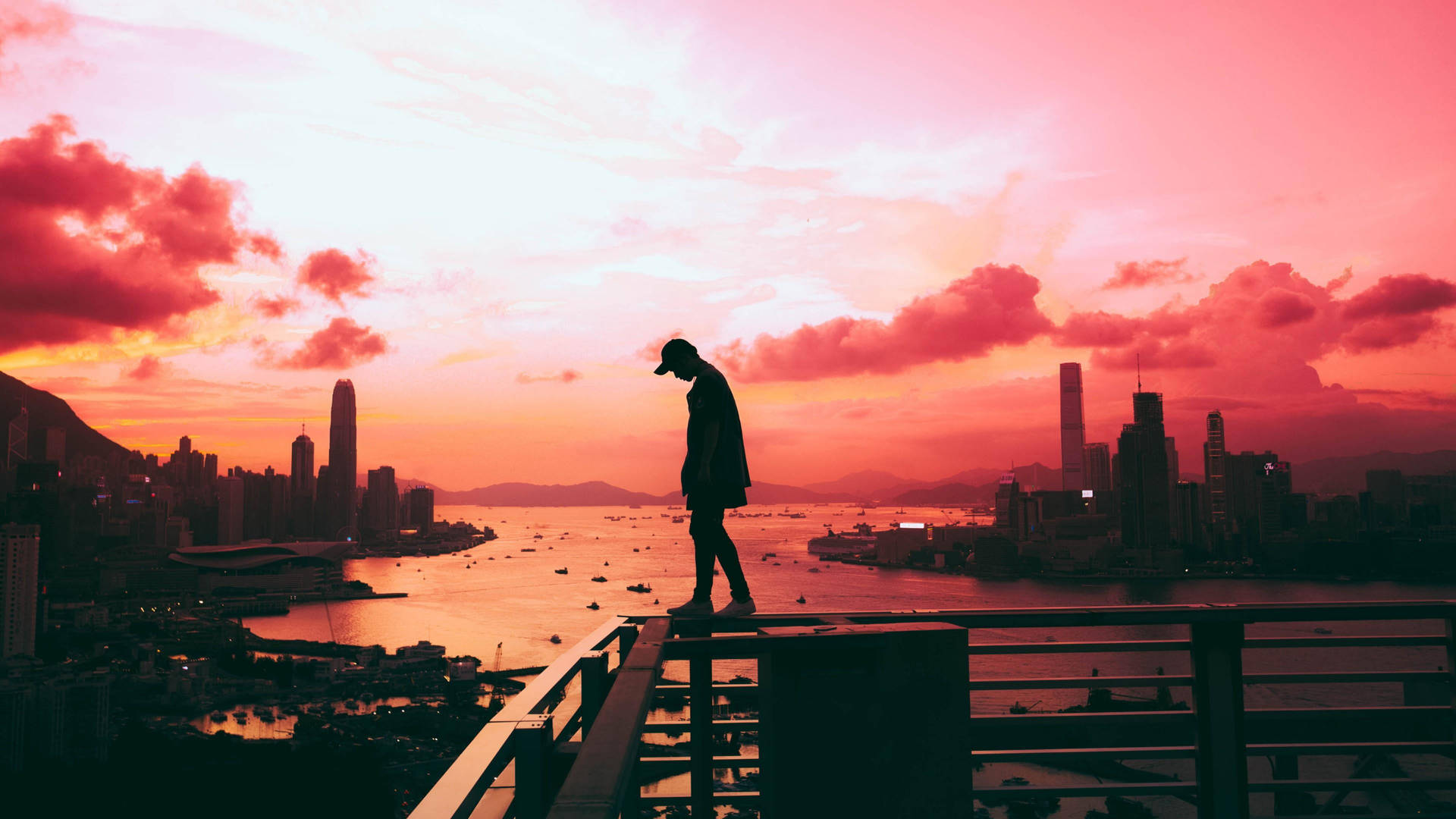 一個人站在欄杆上，觀看繁忙城市和港口上空的彩色日落。