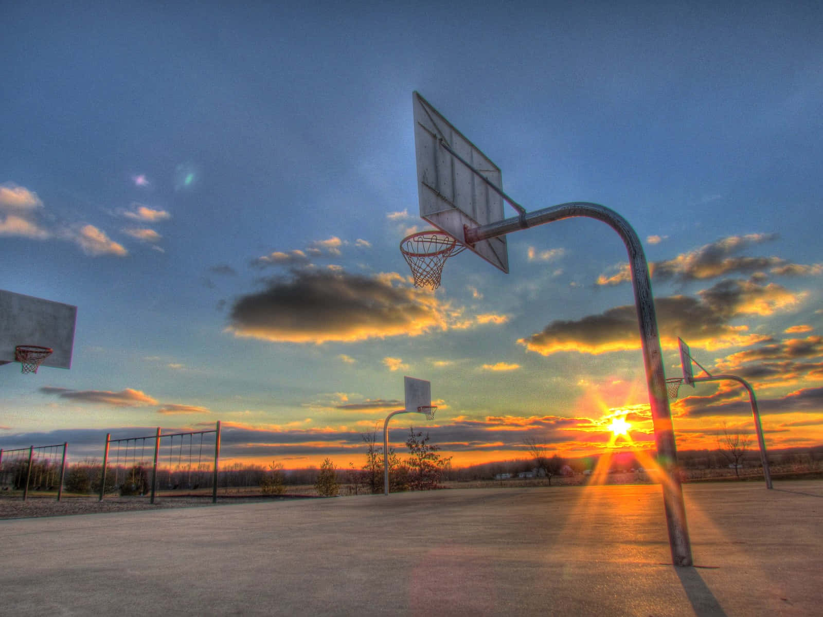 Cool Basketball 桌布 壁紙 — 夕陽下一個風化的籃球場，充滿活力的橙色太陽爆發和戲劇性的雲層投下長長的陰影。