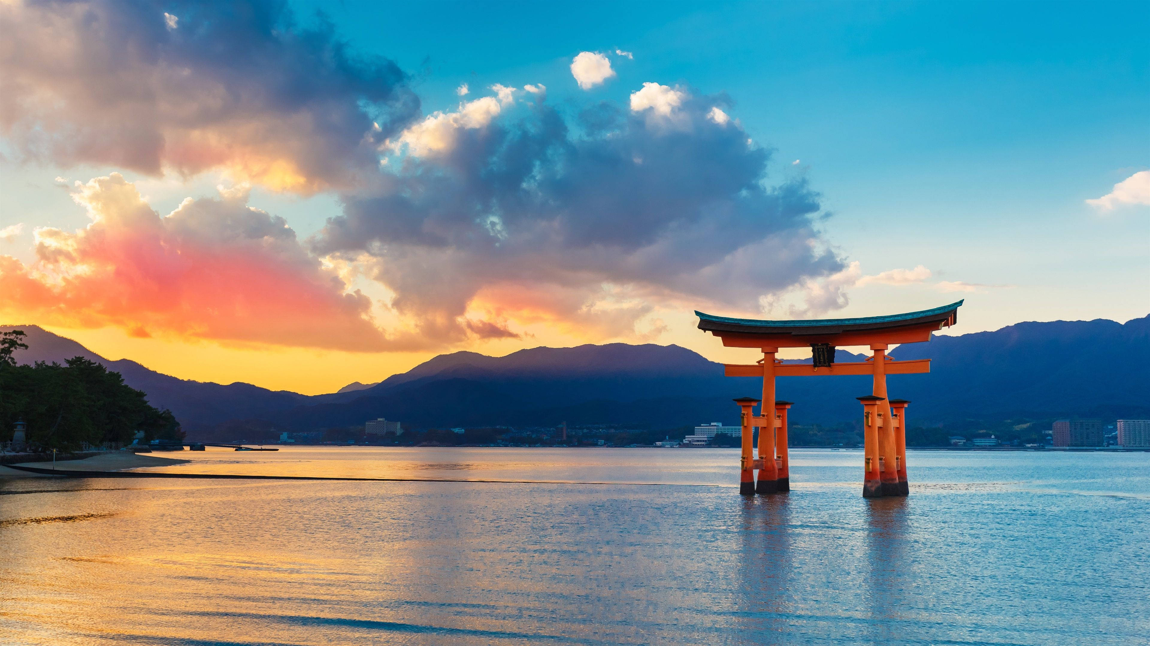 夕陽下的傳統鳥居門 壁紙 — 一座充滿活力的橙色鳥居矗立在日落時分的平靜水面上，背景有山脈和色彩繽紛的雲彩。