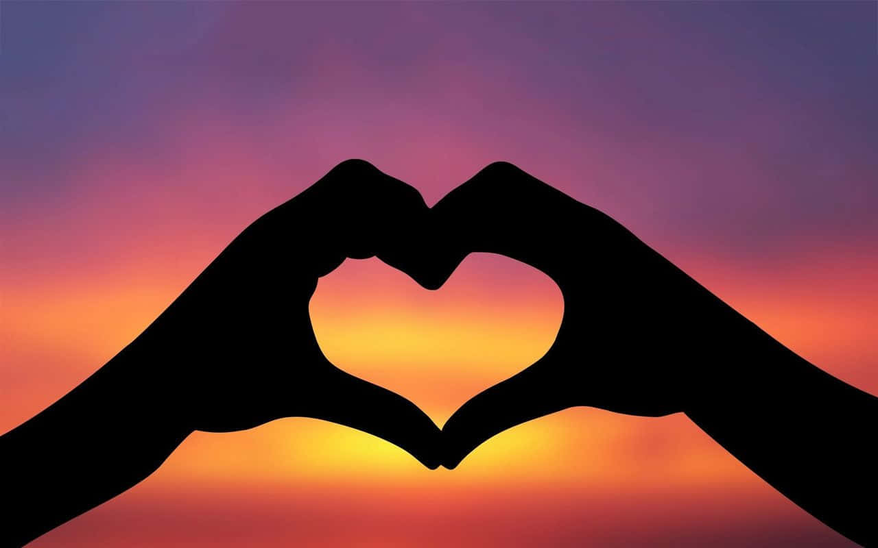 夕陽下的愛意手勢圖片 壁紙 — 剪影的手在融合紫色、橙色和黃色色調的鮮豔日落天空下形成一個心形。