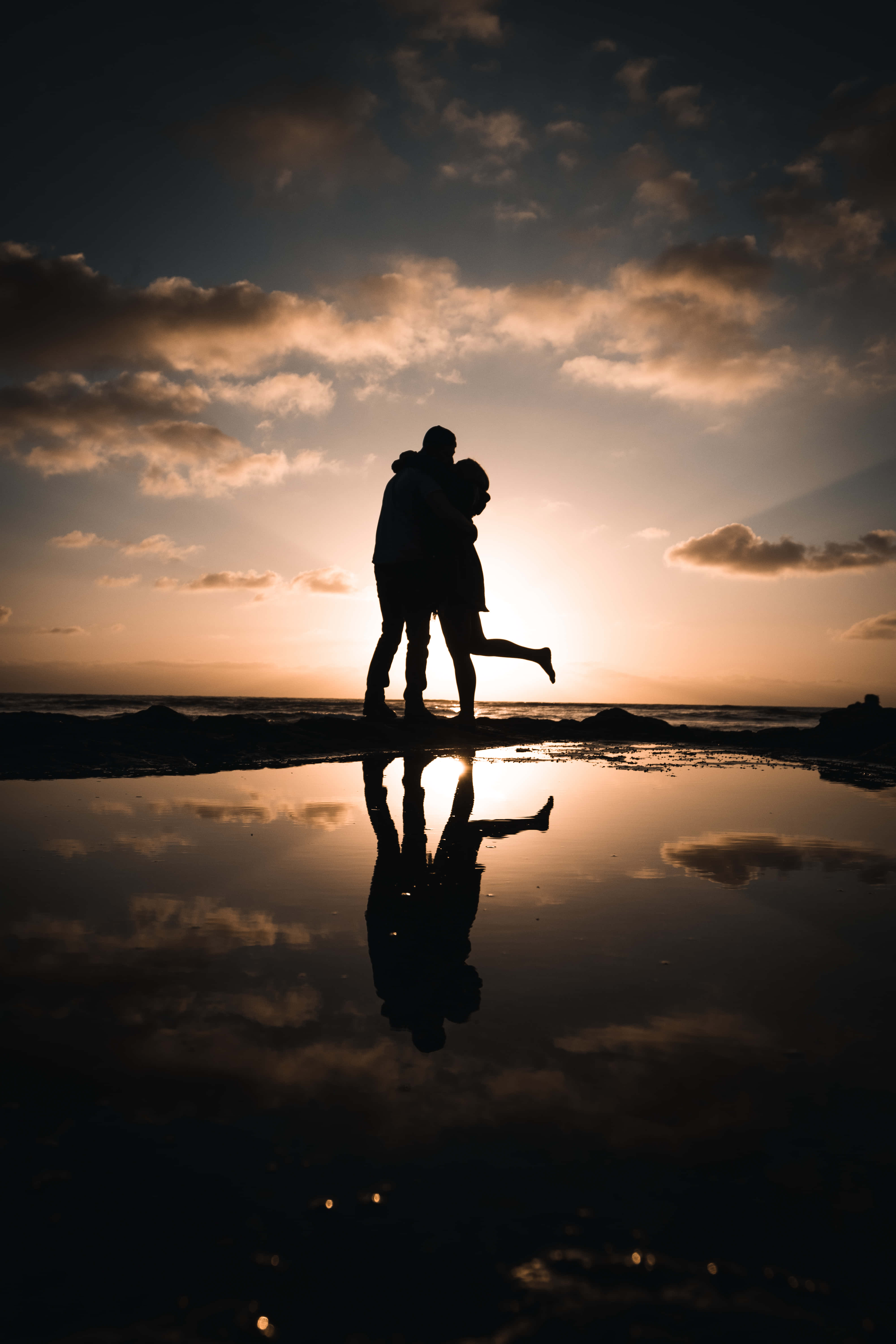 夕陽下親吻情侶照 壁紙 — 一對剪影情侶在日落時分的海灘上擁抱，他們的倒影映照在潮汐池中。