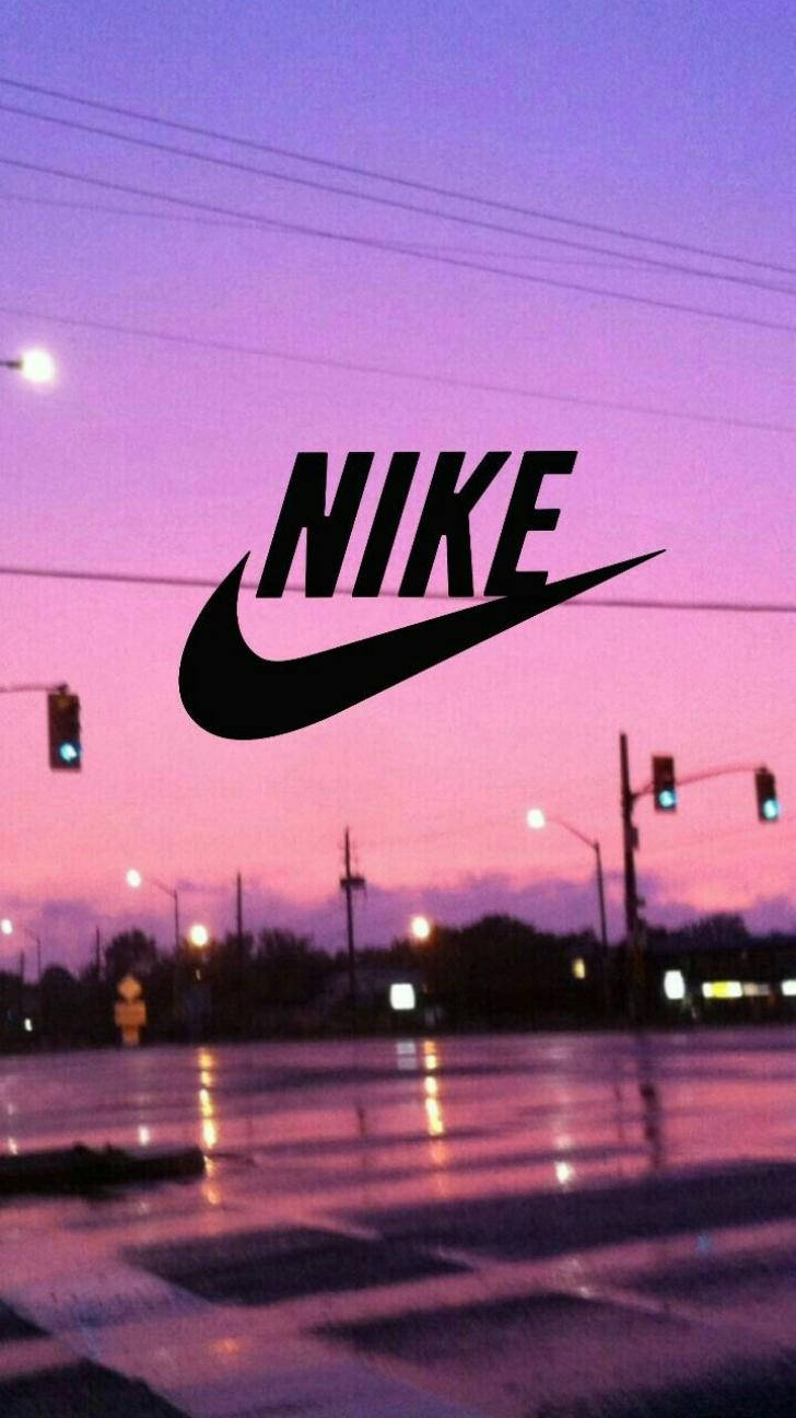 夕陽交叉路口的勾形標誌 壁紙 — 一個黑色Nike標誌漂浮在潮濕的城市街道上，黃昏時分，天空呈現粉紫色，交通燈閃爍發光。