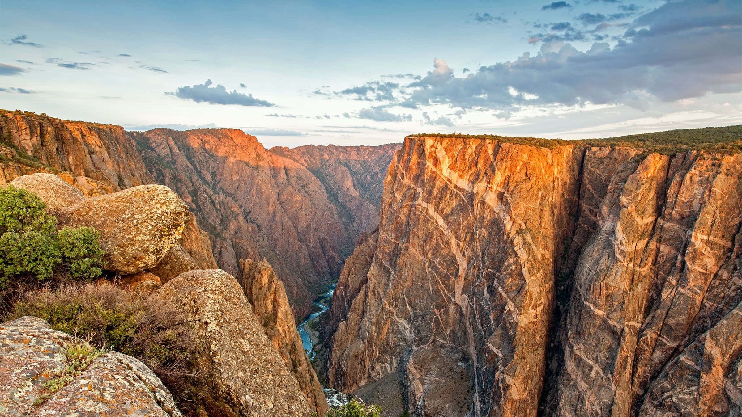 Black Canyon 桌布 壁紙 — 夕陽以溫暖的光芒沐浴廣闊的峽谷，顯露出崎嶇的懸崖和下方蜿蜒的河流。