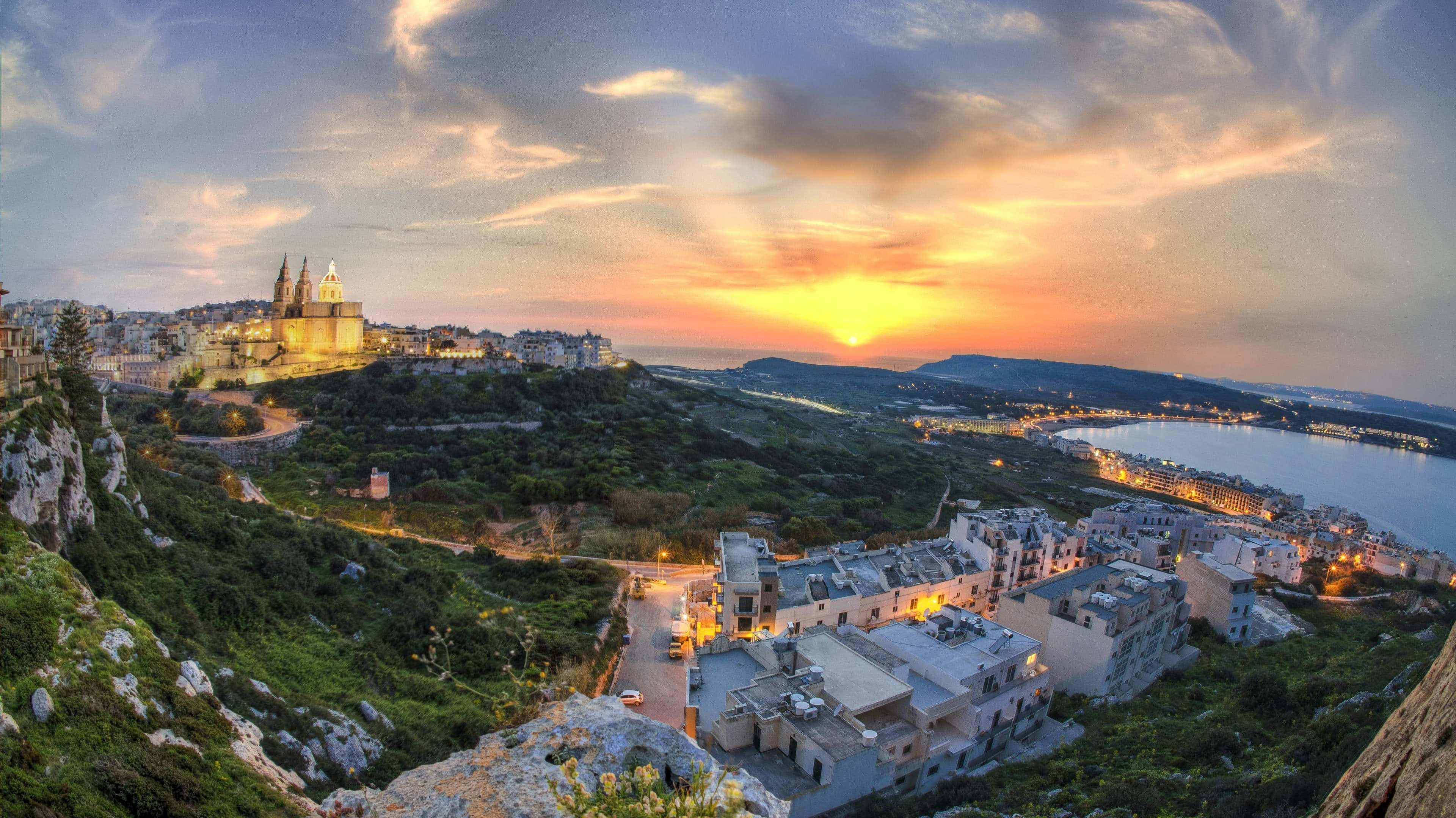 Malta 桌布 壁紙 — 夕陽以溫暖的光芒沐浴懸崖小鎮，古老的石頭建築緊貼在俯瞰平靜大海的綠色山丘上。