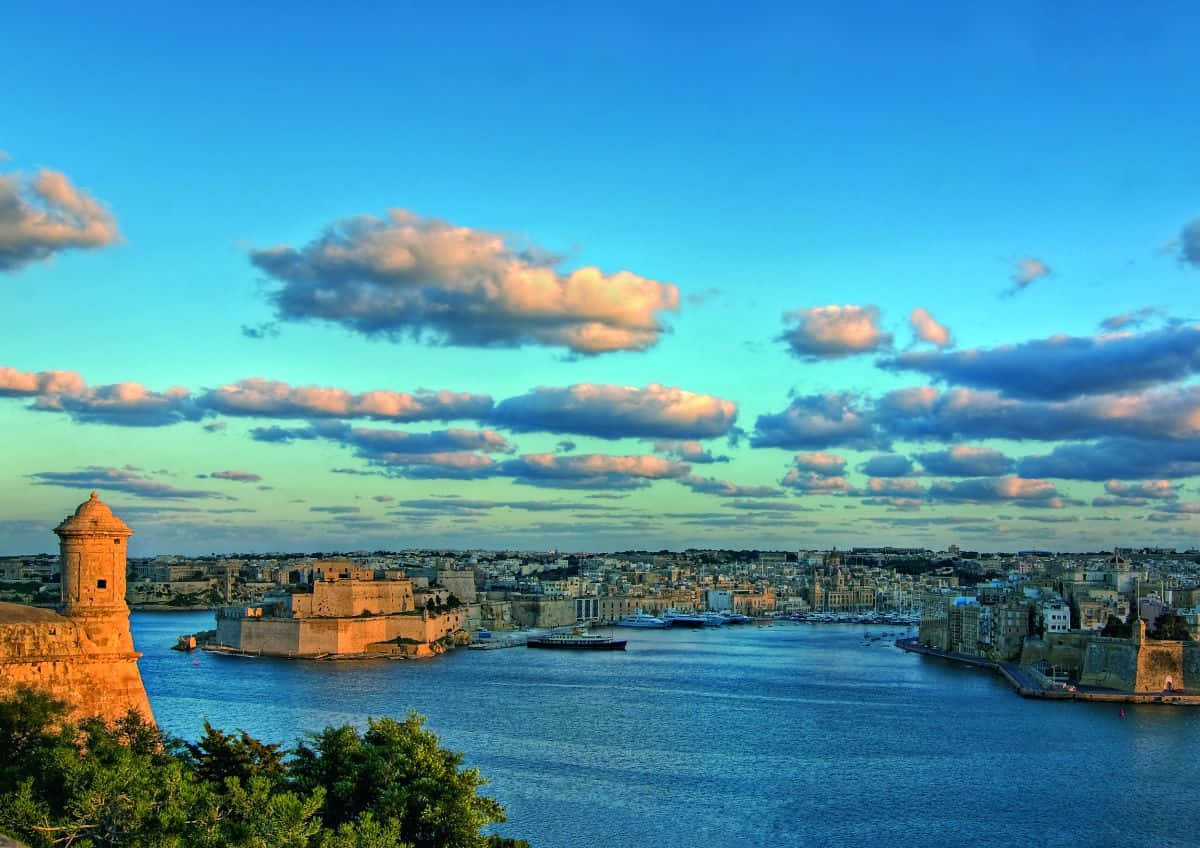 Malta 桌布 壁紙 — 夕陽以溫暖的金色光芒沐浴歷史悠久的沿海堡壘和城市景觀，映襯著帶有散雲的鮮豔藍天。