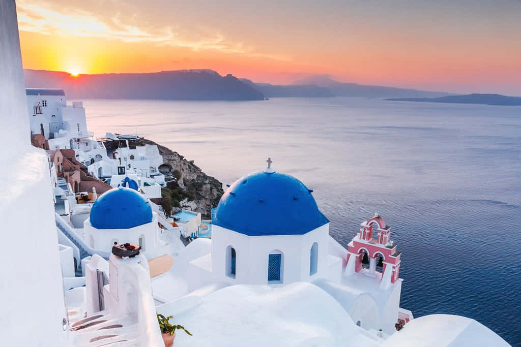 Greek Island 桌布 壁紙 — 夕陽以溫暖色調描繪天空，映照在俯瞰愛琴海的懸崖上那些標誌性的藍頂教堂之上。
