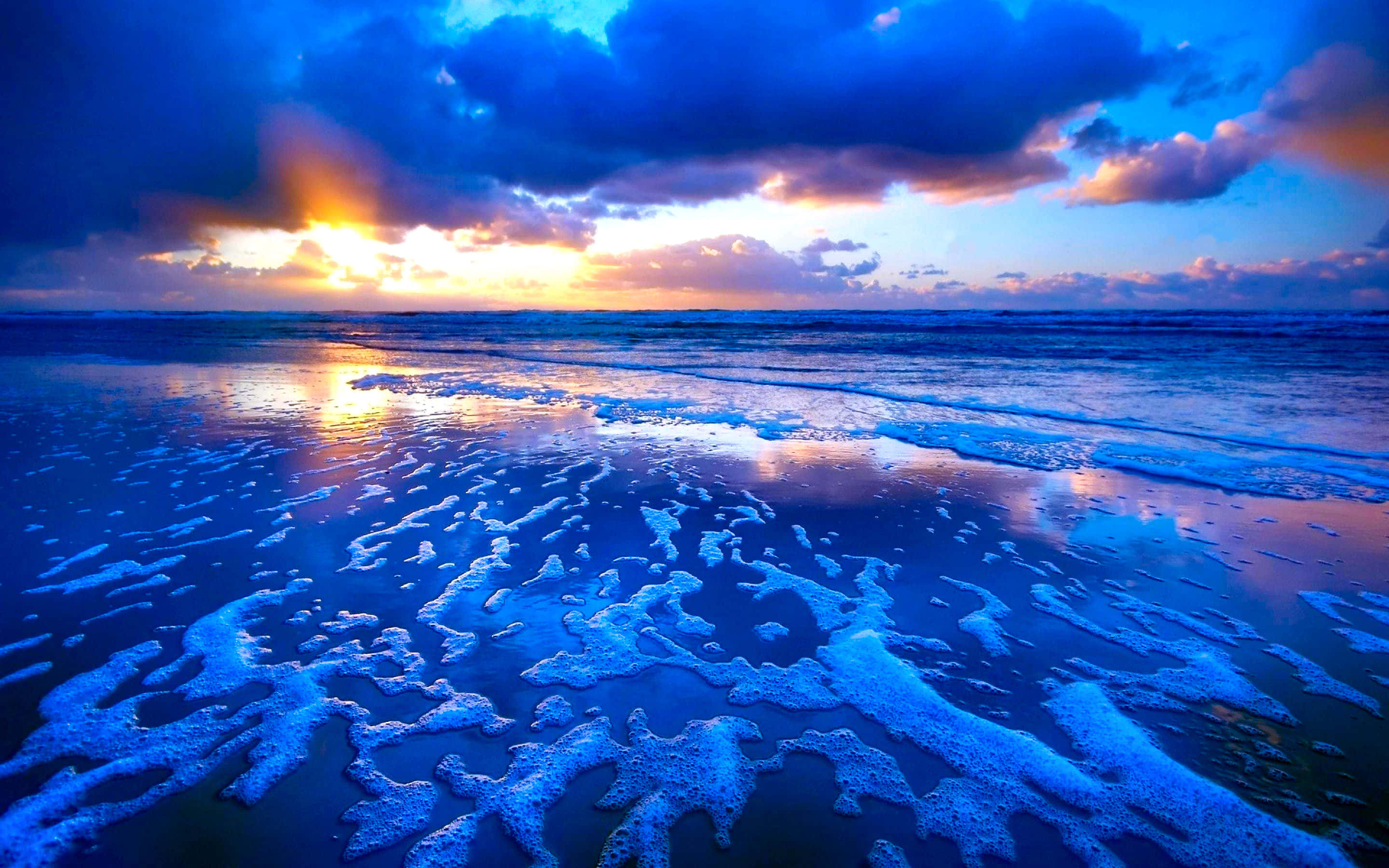 Horizon 桌布 壁紙 — 夕陽以火橙色和深藍色描繪海洋，海浪在戲劇性的雲層下拍打泡沫覆蓋的海岸。
