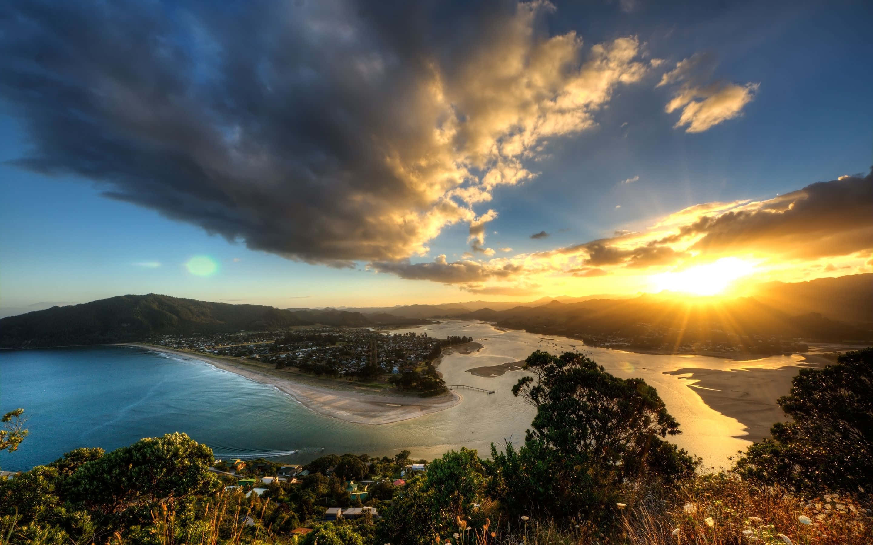 New Zealand 桌布 壁紙 — 夕陽以金色光芒沐浴著海岸灣，戲劇性的雲朵在蜿蜒的海岸線和遠處的山丘上空盤旋。