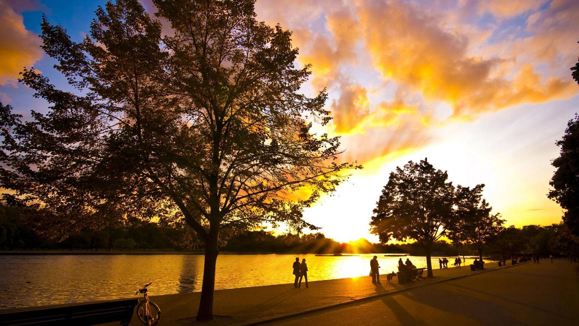 夕陽公園風景 壁紙 — 剪影人物沿湖邊小徑漫步放鬆，金色夕陽光芒穿過戲劇性的雲層迸發而出。