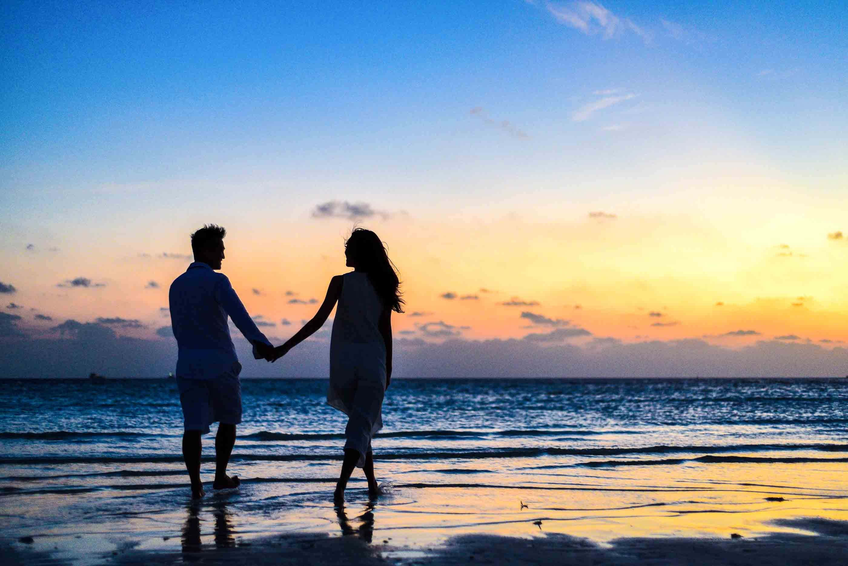 夕陽剪影戀愛模範照片 壁紙 — 一對情侶手牽手沿著濕漉漉的海灘在日落時分漫步，他們的剪影在漸變天空下的水中倒映。