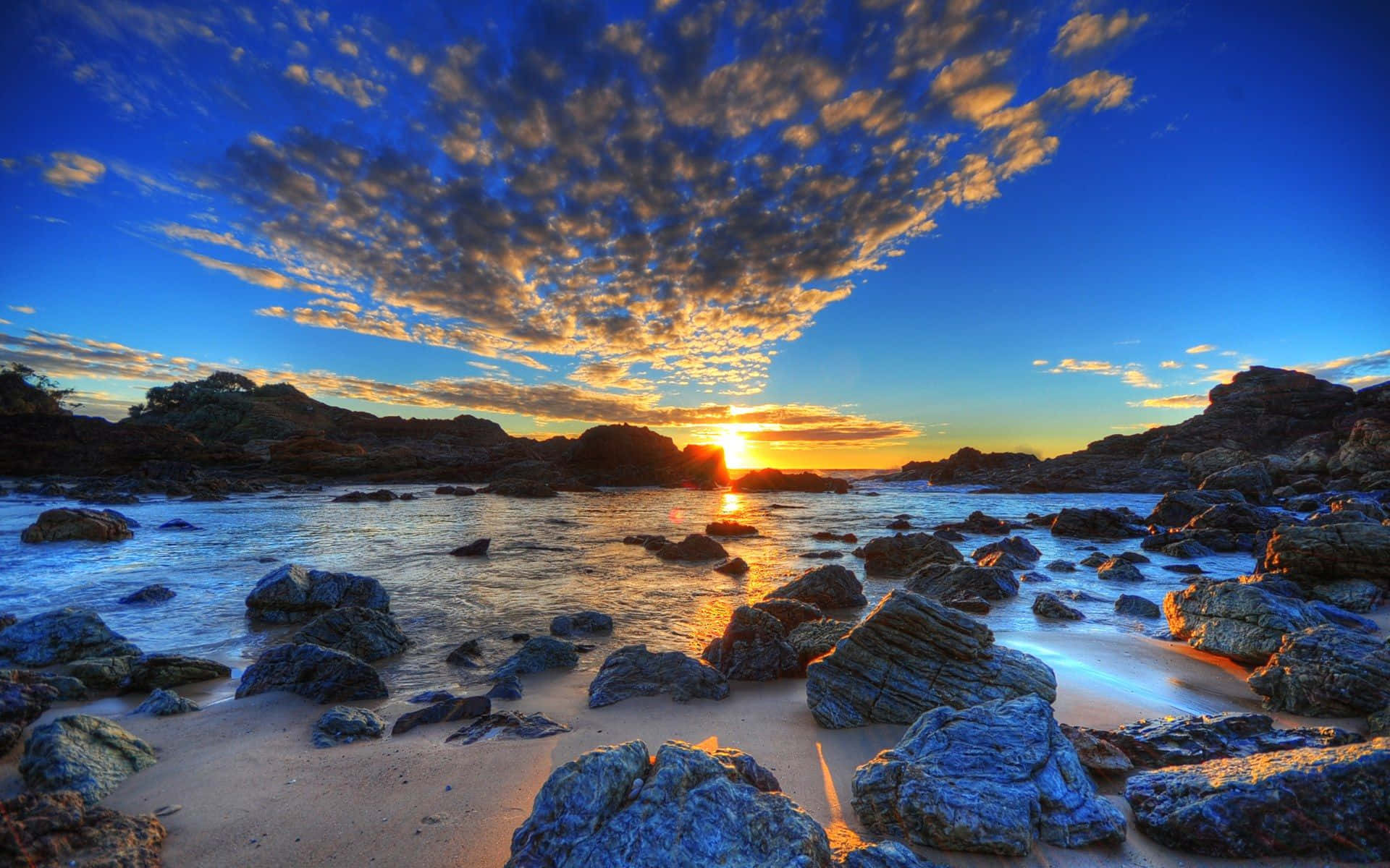 Australia Beach 桌布 壁紙 — 夕陽在岩石海灘上繪出金色雲彩，海浪輕輕拍打石頭和沙灘。