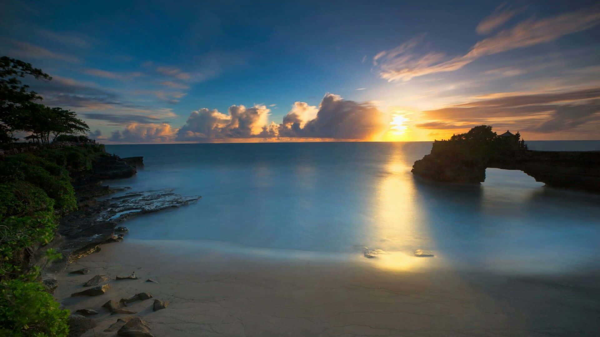 Bali Beach 桌布 壁紙 — 夕陽在海面上閃耀，金色的光芒映照在平靜的水面上，旁邊是岩石海岸和茂密的綠植。