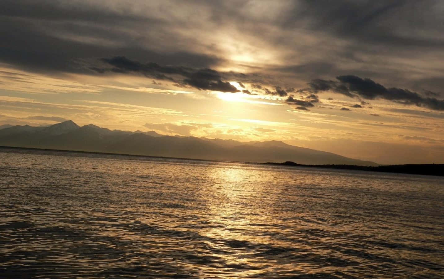 Sunset Reflection 桌布 壁紙 — 夕陽在漣漪的水面上閃耀金光，遠山在戲劇性的雲層下形成剪影。