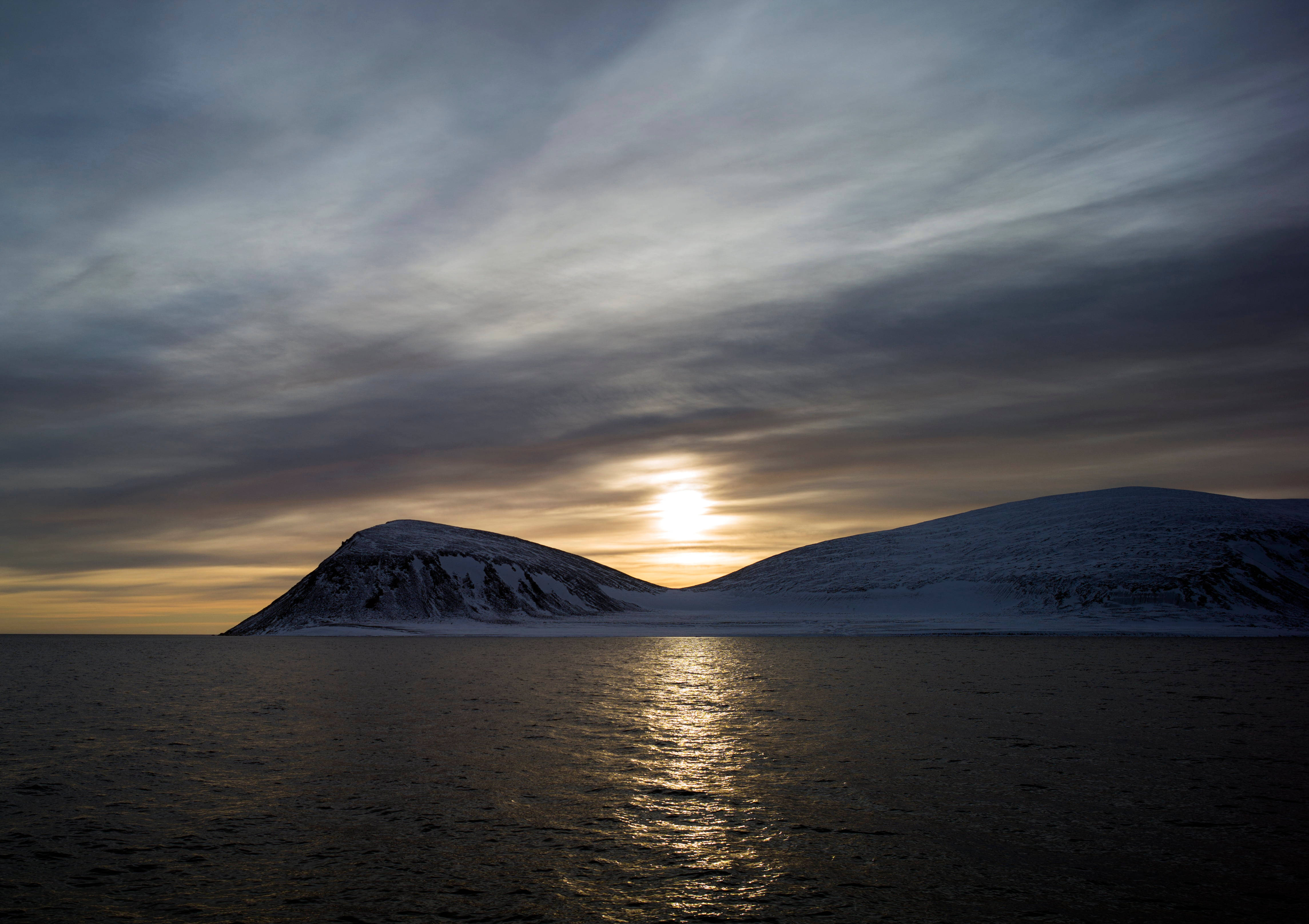 Svalbard 桌布 壁紙 — 夕陽在積雪的山峰後溫暖地發光，映照在條紋雲下平靜的水面上。