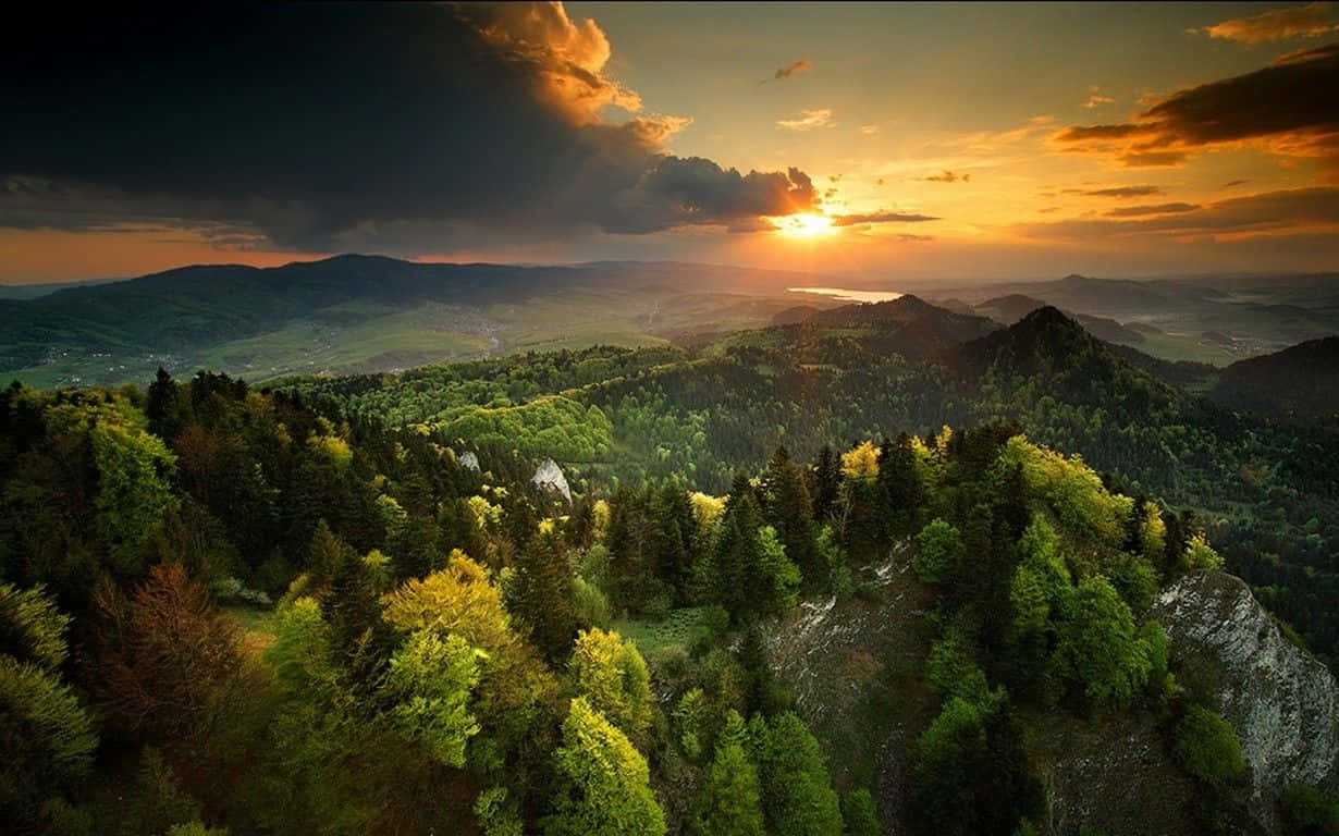 Spring Sunset 桌布 壁紙 — 夕陽在連綿的綠色山丘和茂密的森林上繪出戲劇性的天空，遠山隱約可見。