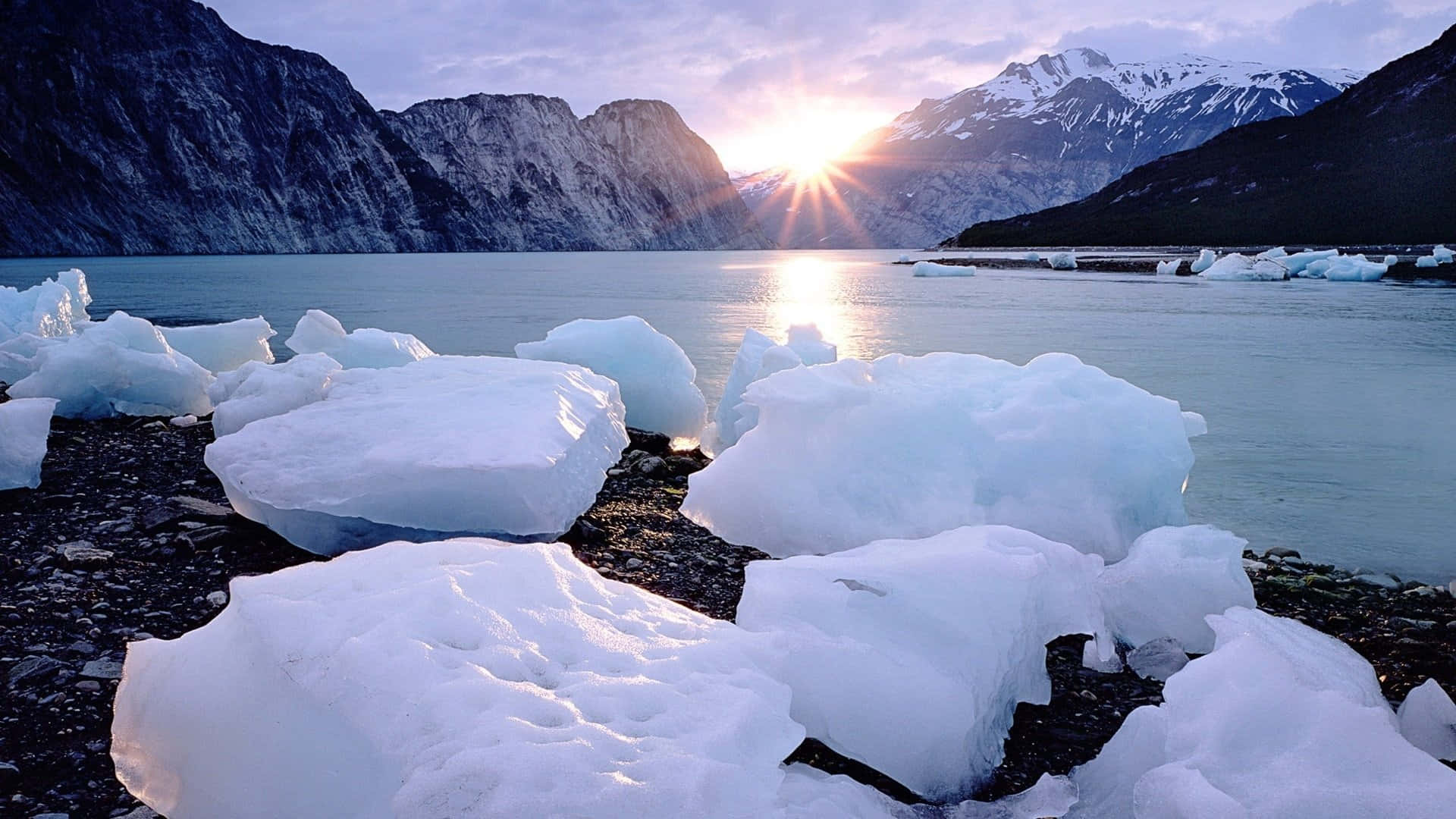 Iceberg 桌布 壁紙 — 夕陽在雪山後方閃耀，將金色光芒投射在平靜冰川湖上漂浮的冰山上。