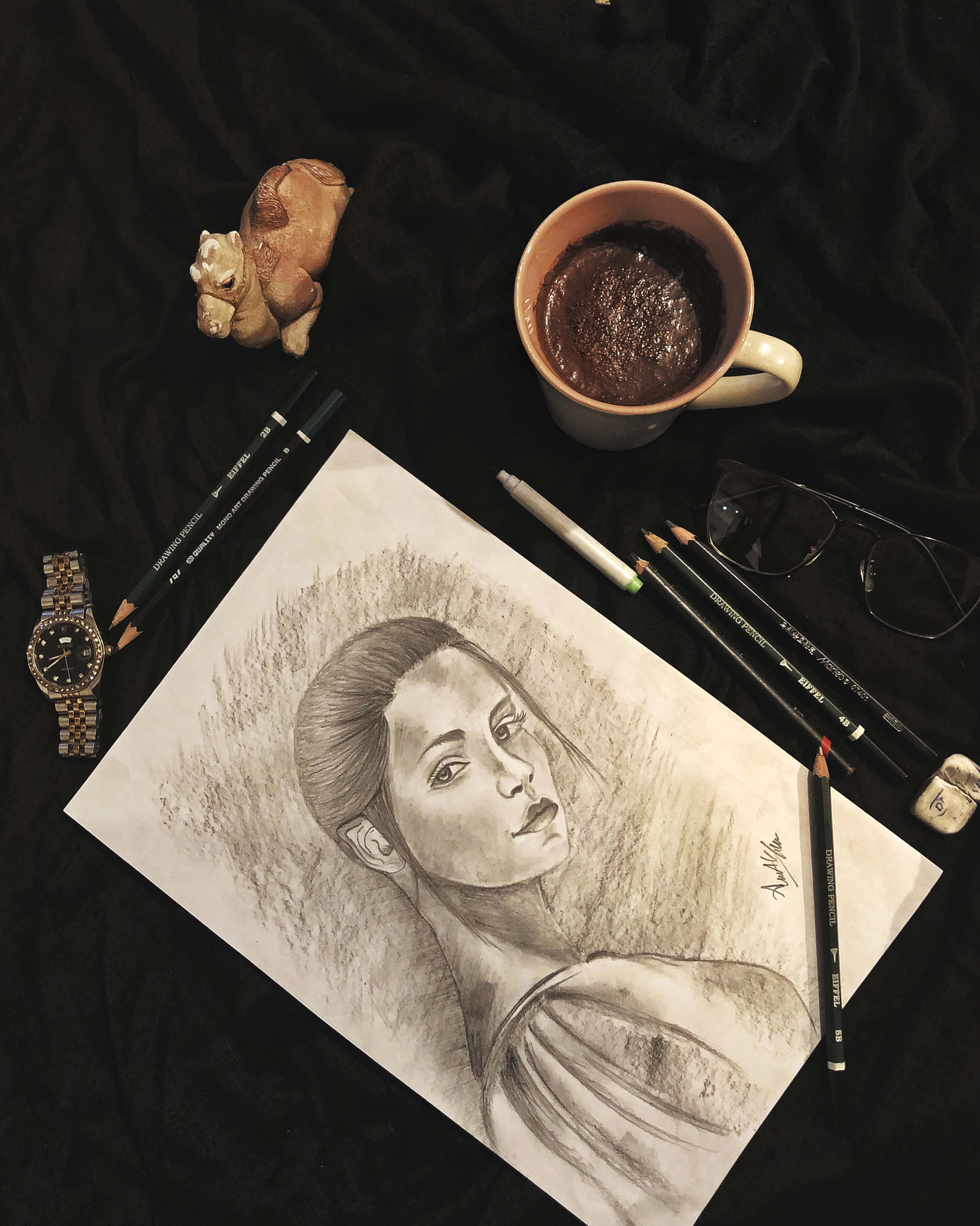 夕陽夢境：飄浮熱氣球之旅 壁紙 — 一幅詳細的鉛筆素描女性肖像畫放在深色布料上，周圍環繞著美術用品和一杯咖啡。